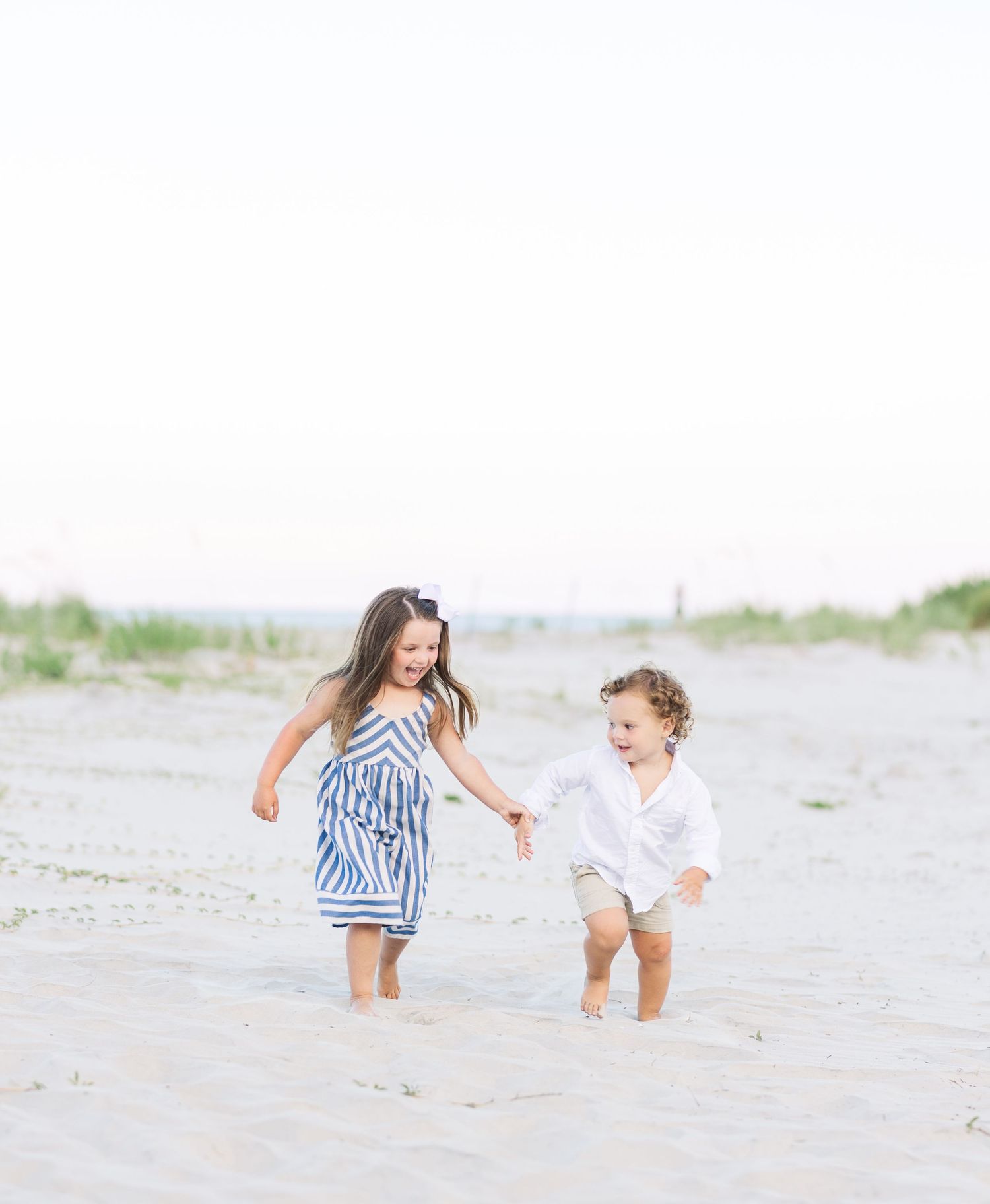 Two children play and run together on a sandy beach wearing summer clothes.