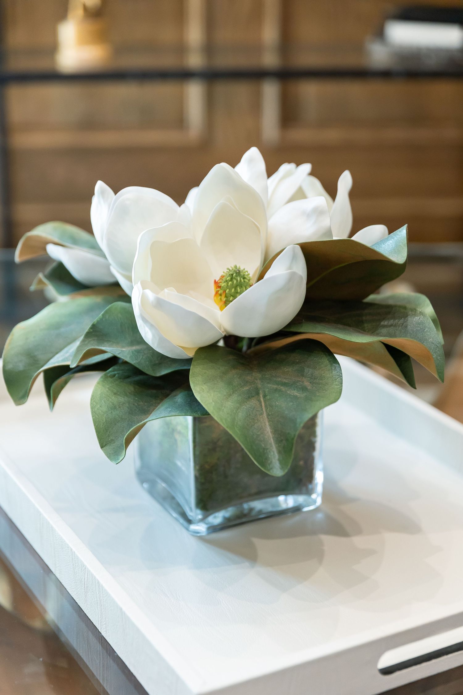 Single white magnolia bloom with green leaves arranged in square glass vase on white tray.