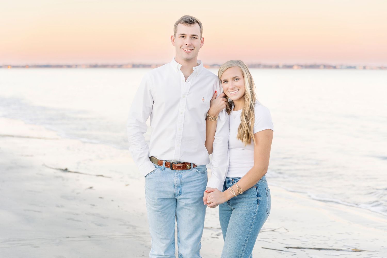 Couple in matching white tops and jeans posing together on a beach during sunset.