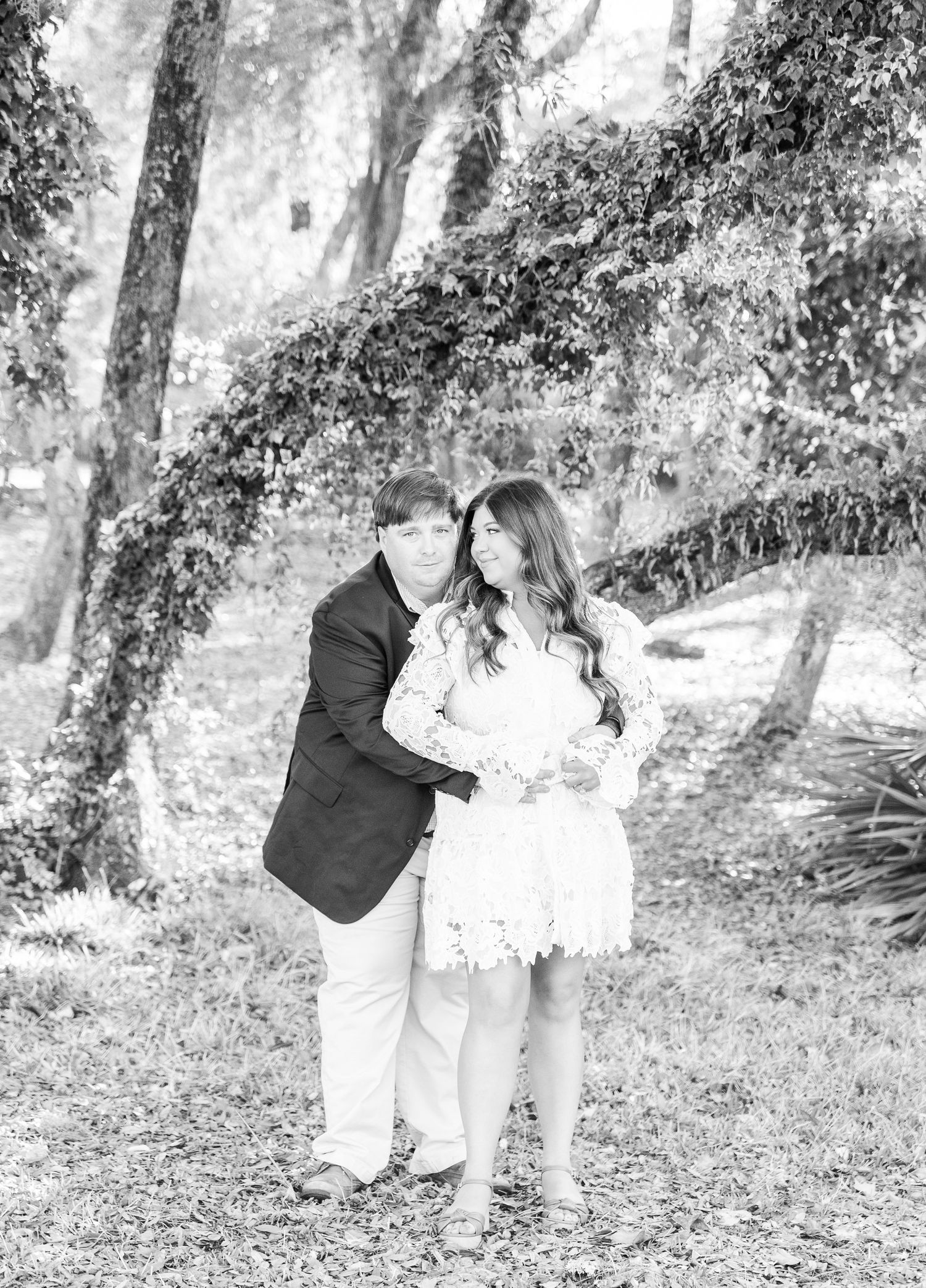 A romantic black and white photo of a couple embracing in a wooded setting.