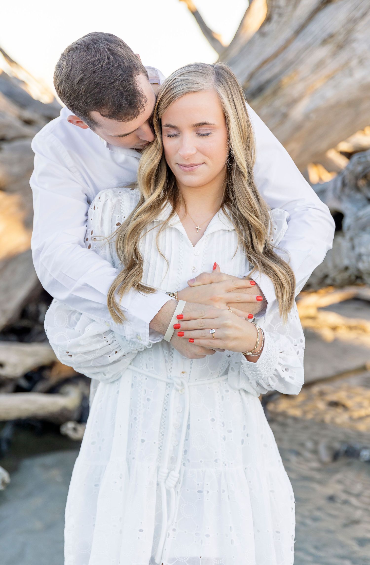 An intimate embrace captured on a beach with driftwood and soft lighting.