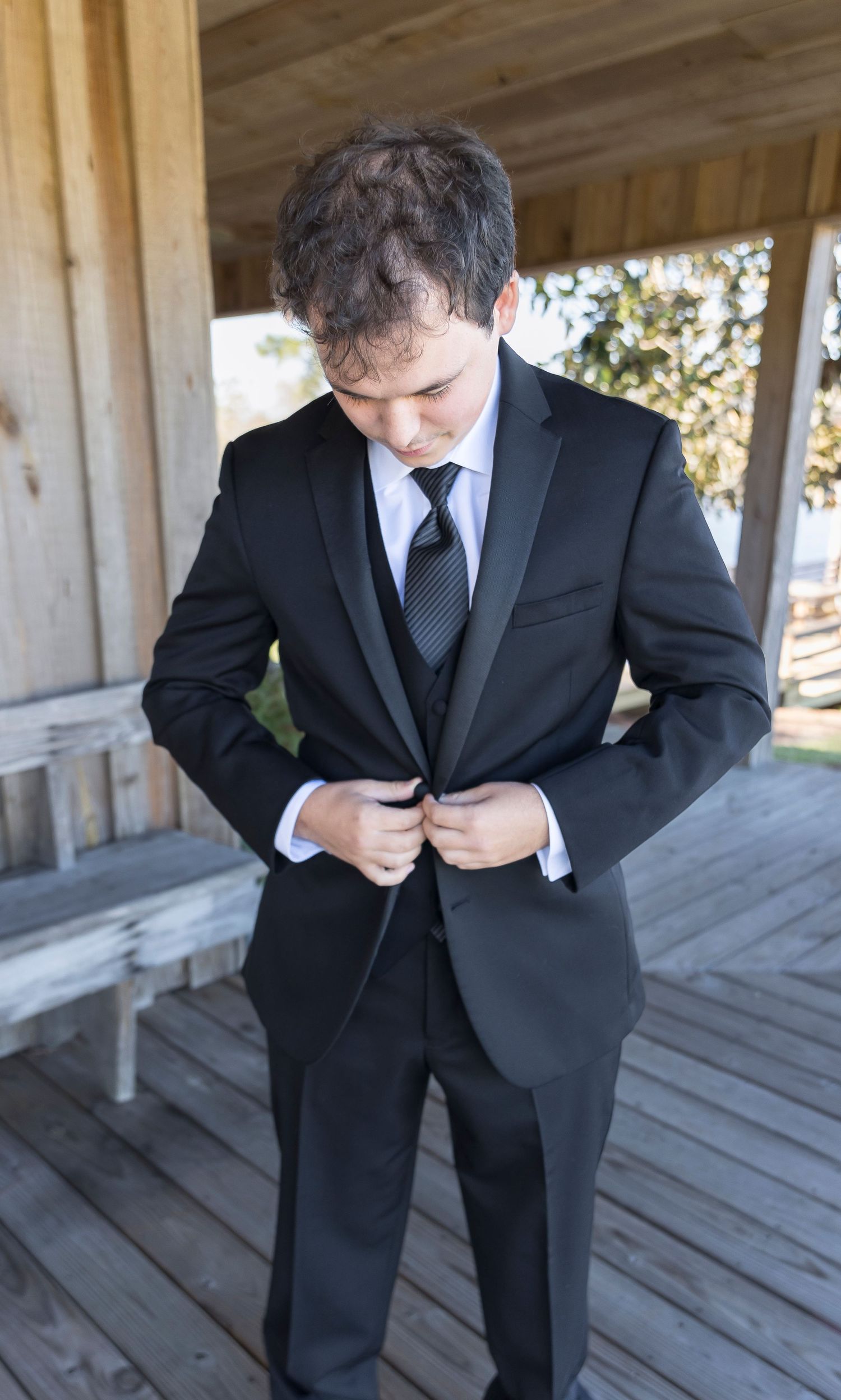 Person in black suit adjusts jacket while standing on wooden porch.