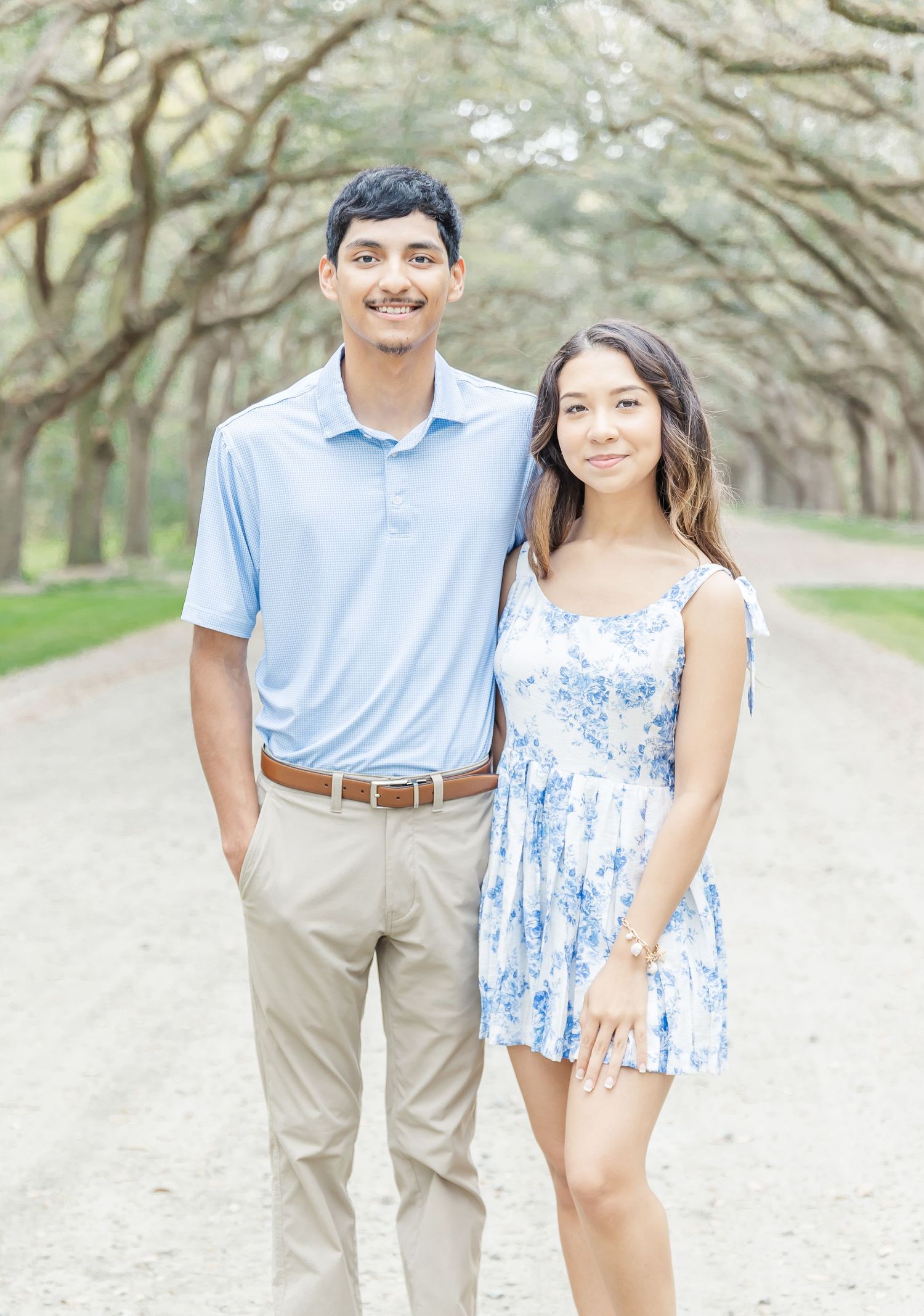 Young couple in coordinated blue and khaki outfits pose on oak-lined pathway.