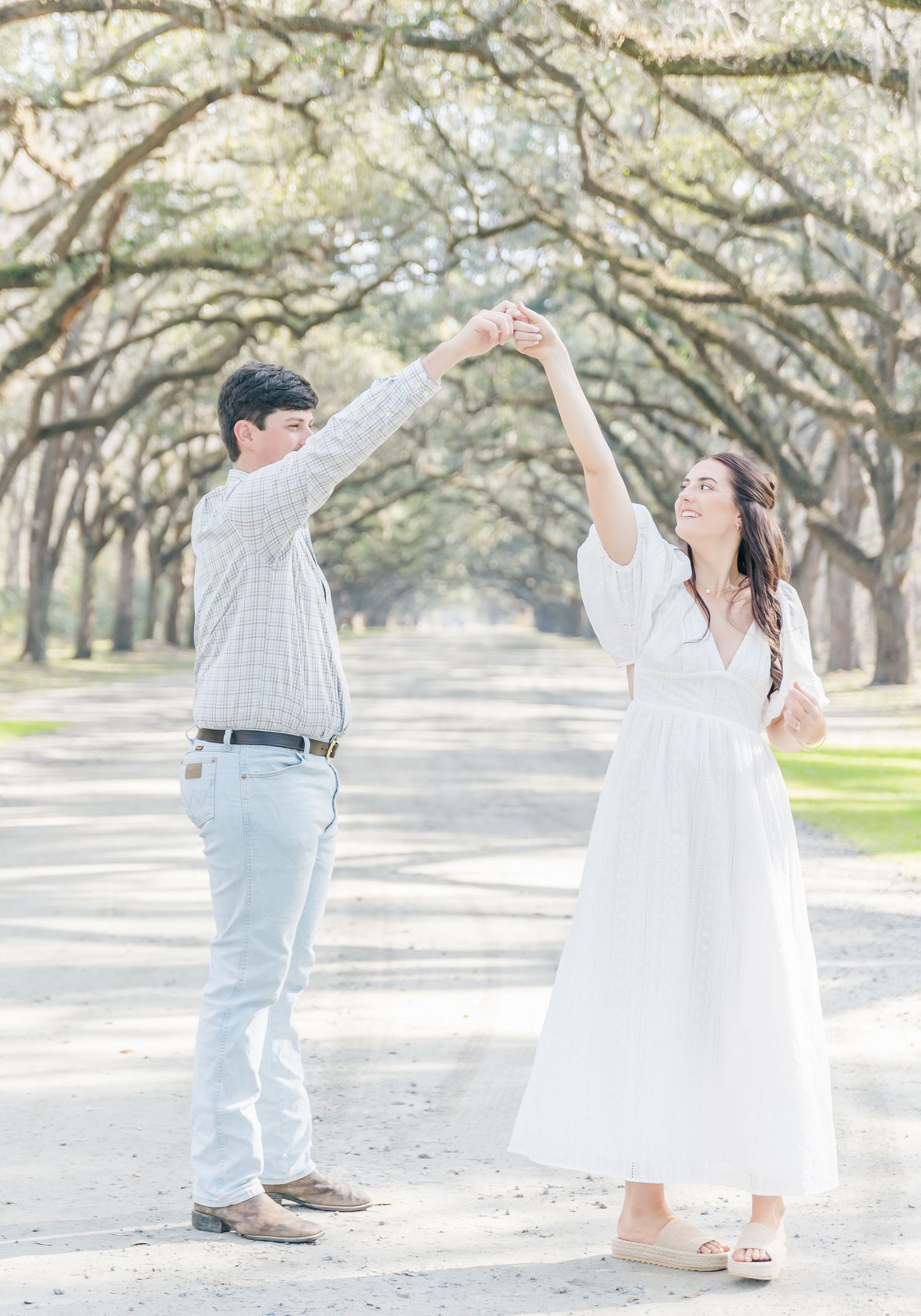 A couple share a playful dance moment beneath an archway of oak trees.