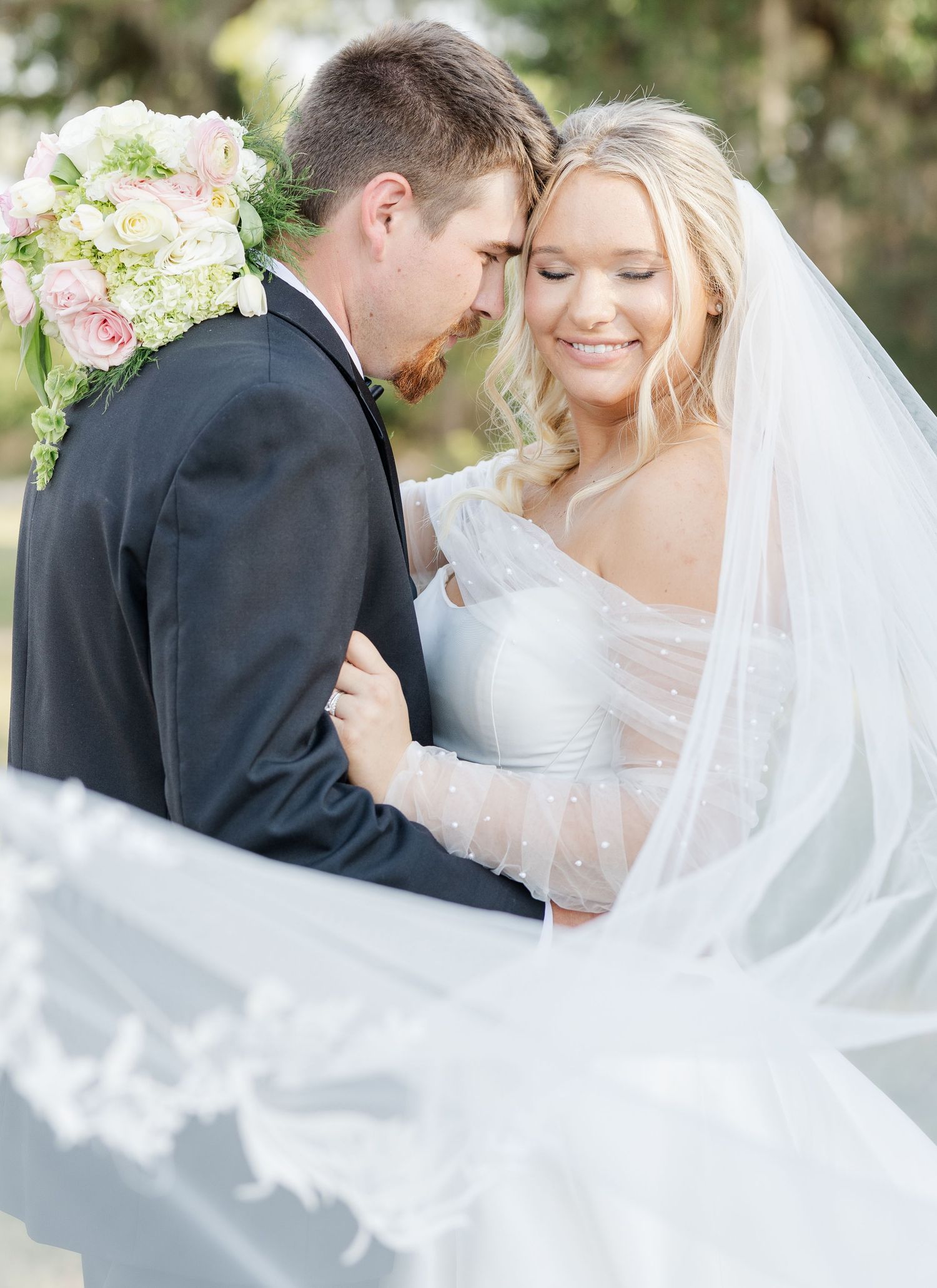 A bride in a flowing veil smiles as she embraces her groom in a dark suit, holding a pink floral bouquet.