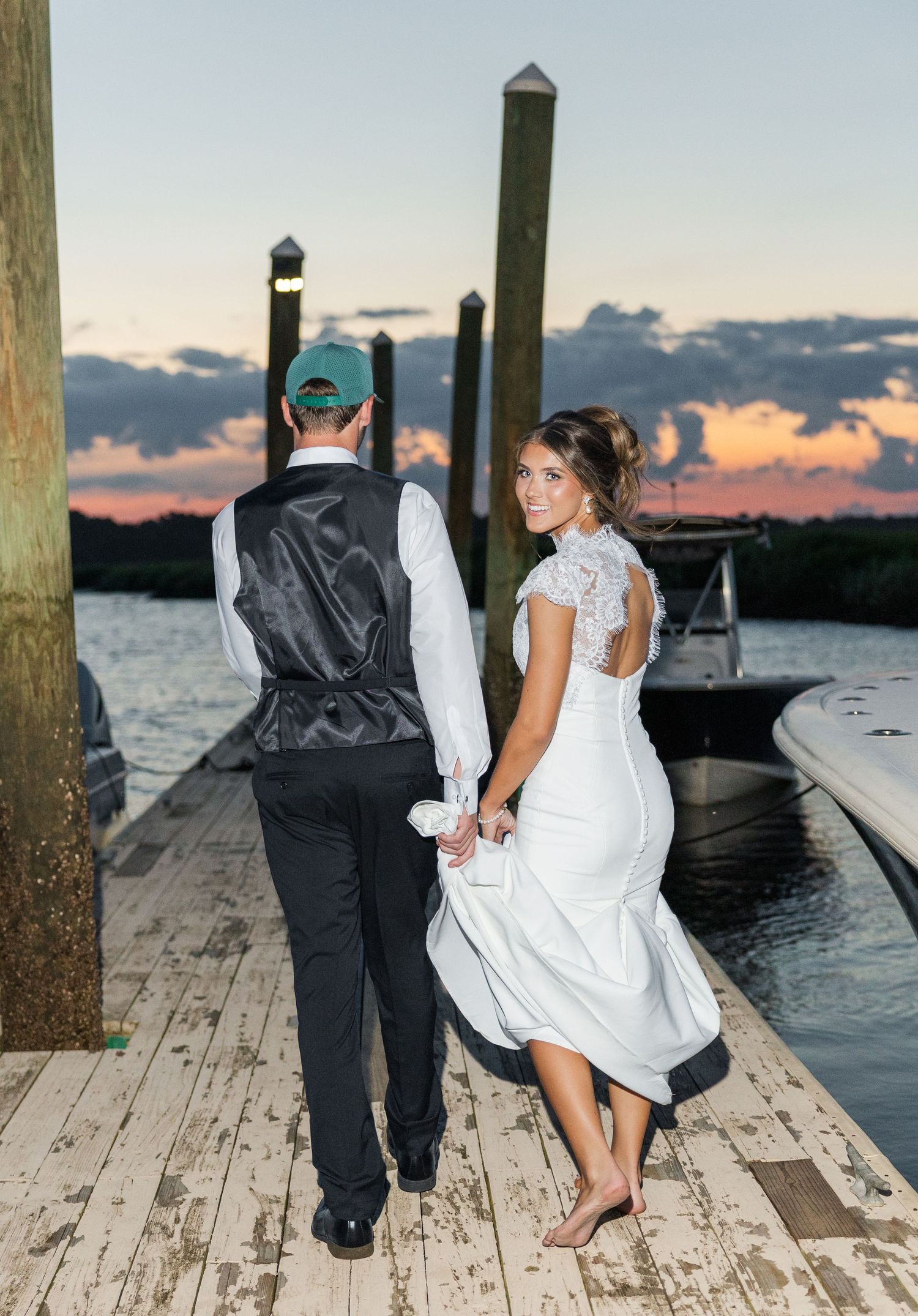 Bride and groom holding hands on a wooden dock at sunset, bride in lace back wedding dress, groom in vest and cap.