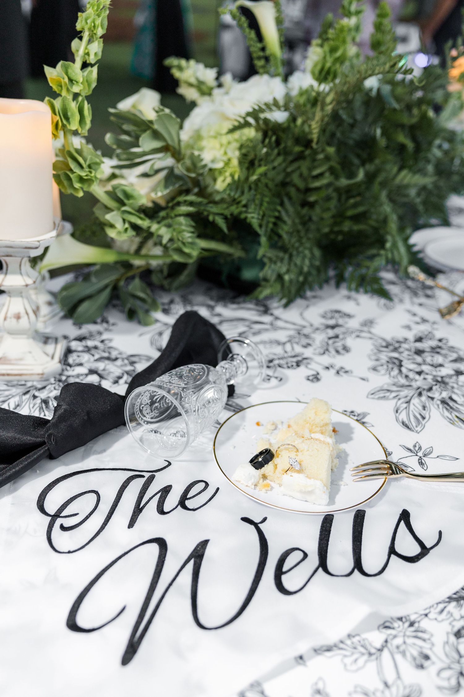 Elegant wedding sweetheart table with The Wells sign, black toile tablecloth, white floral centerpiece, and slice of wedding cake.