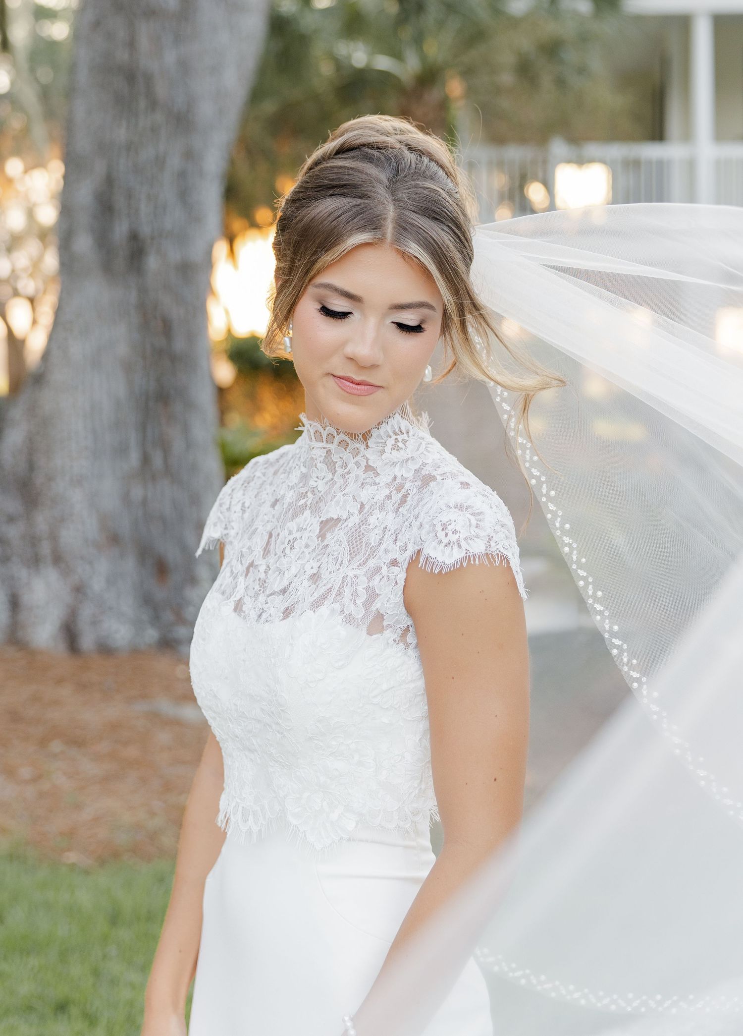 Bride in lace high-neck wedding gown with flowing veil, elegant updo and soft makeup, posing outdoors.