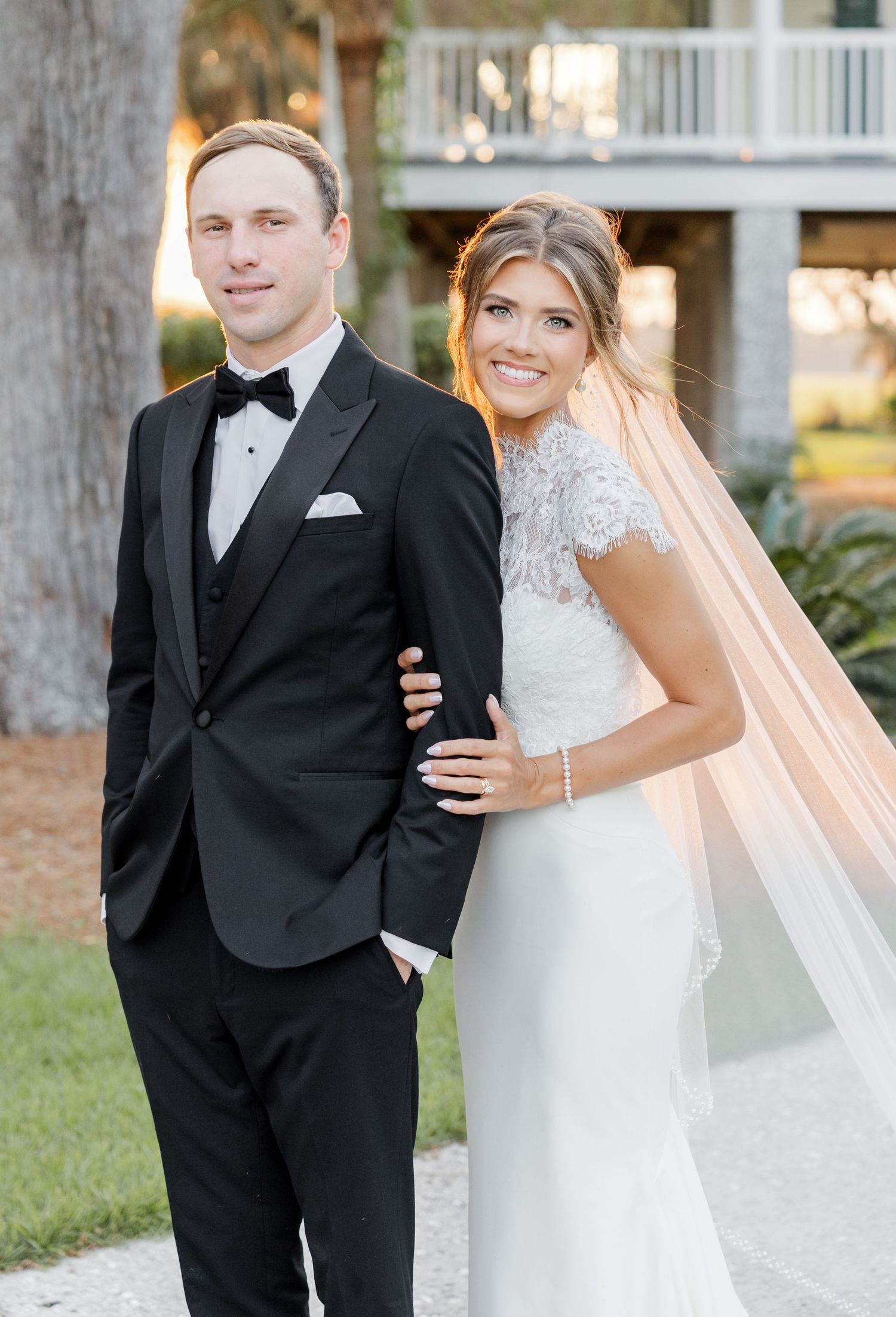 Bride in lace gown and veil poses with groom in black tuxedo outdoors at elegant wedding venue.