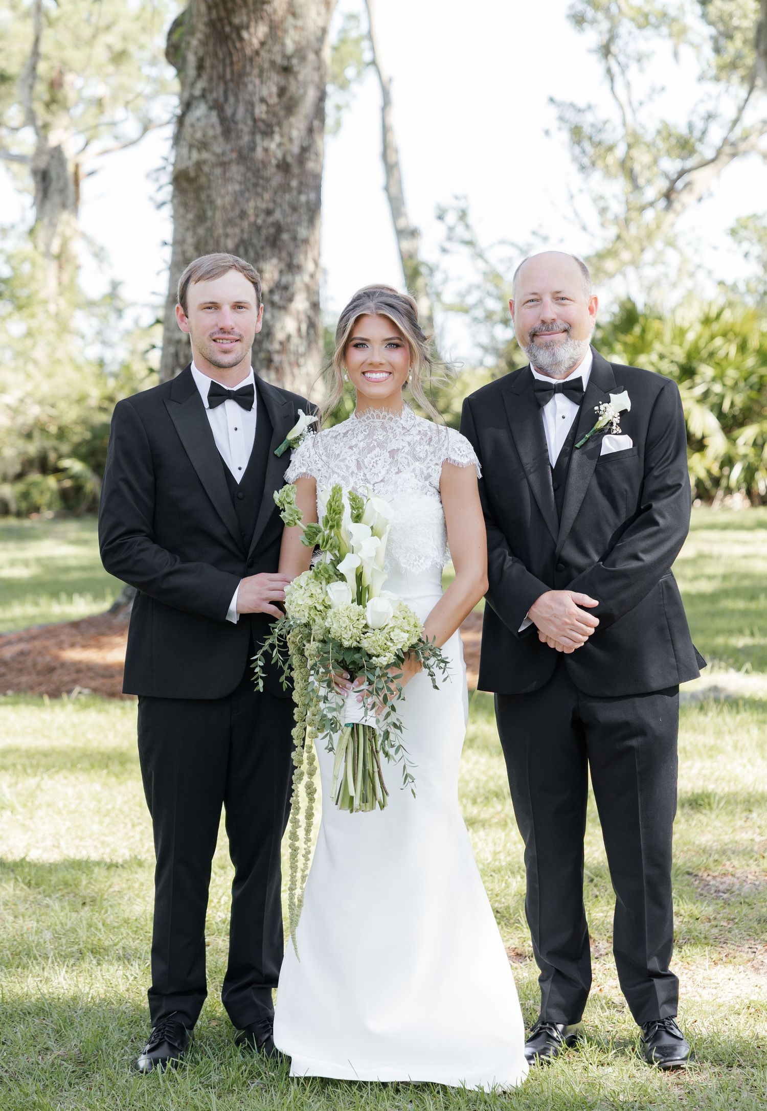 Bride in lace gown holding bouquet poses with two men in black tuxedos outdoors at a wedding.