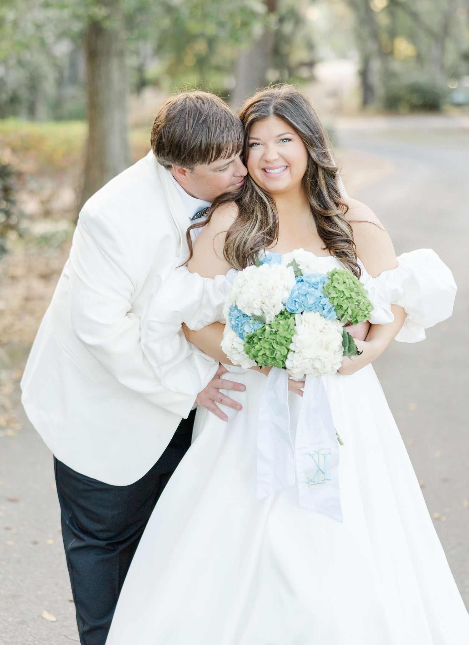 Bride smiling holding blue and white hydrangea bouquet while groom in white jacket kisses her cheek outdoors.