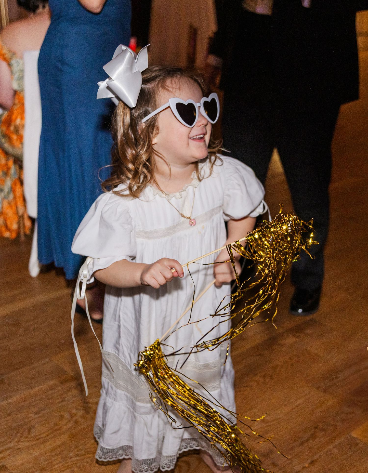 Adorable flower girl wearing heart sunglasses dances with gold tinsel streamers at wedding reception.