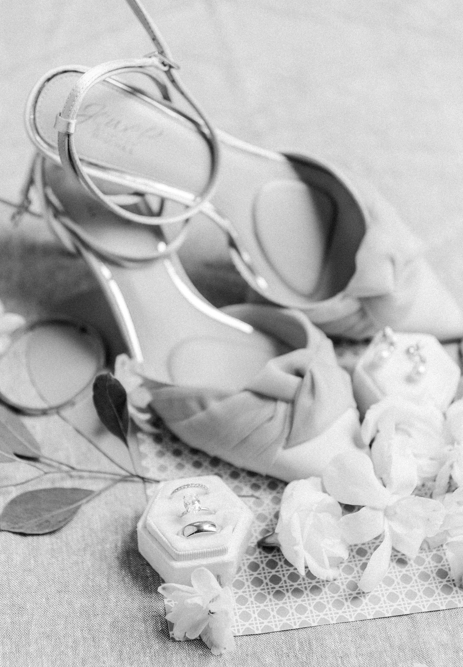 Black and white photo of blue bow heels, wedding rings in hexagon box, and white floral details.