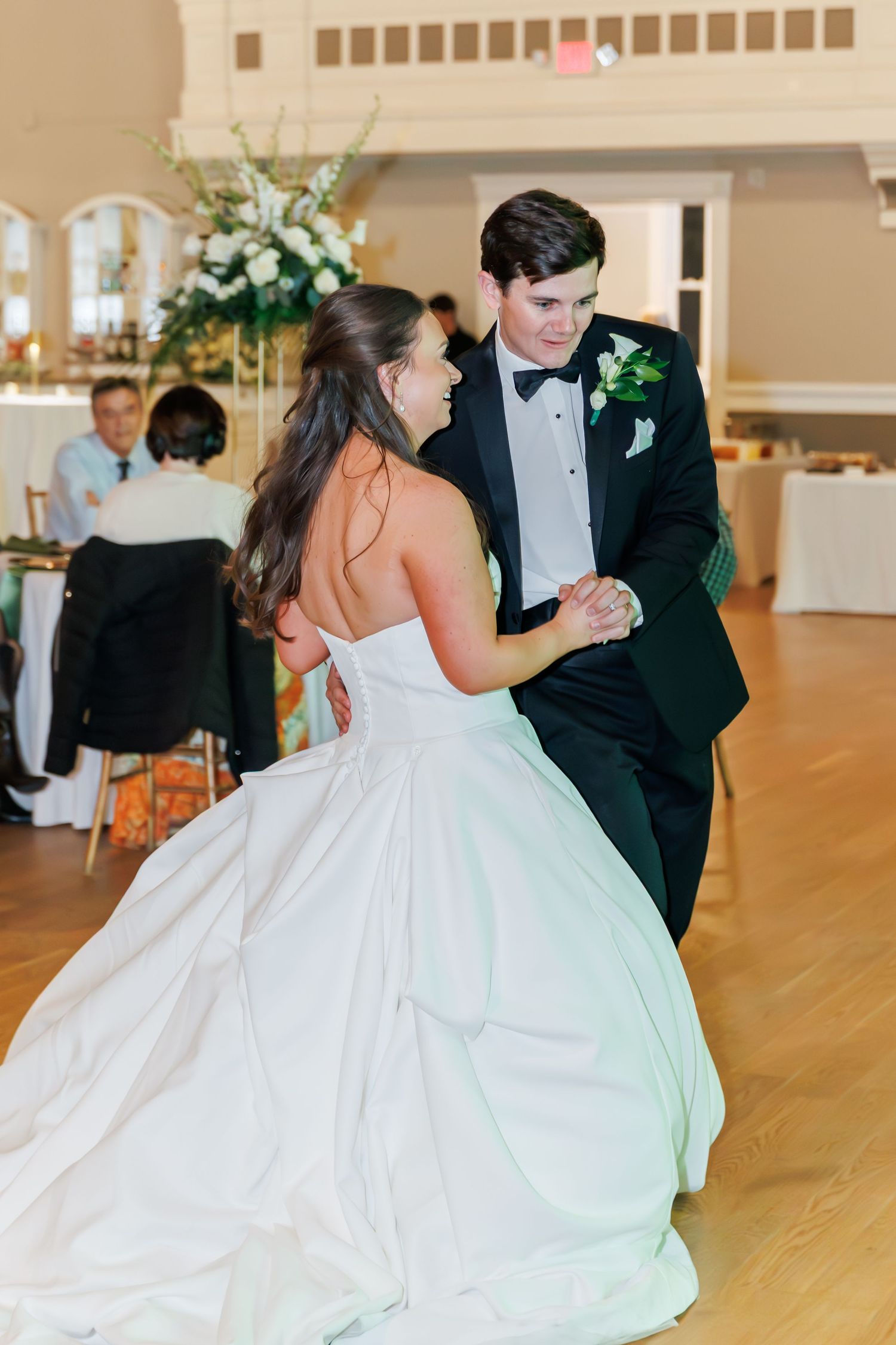 Bride and groom share their first dance at wedding reception, bride in strapless white gown, groom in black tuxedo.