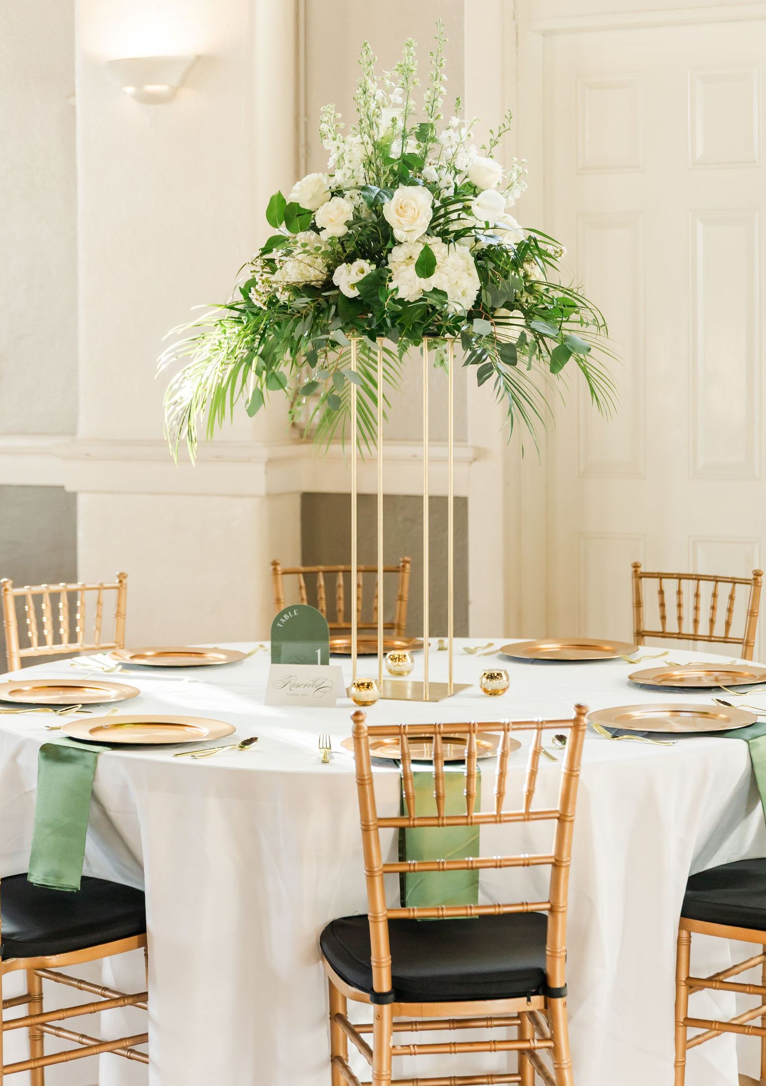 Tall gold stand centerpiece with white roses and tropical greenery on white tablecloth with gold chiavari chairs.