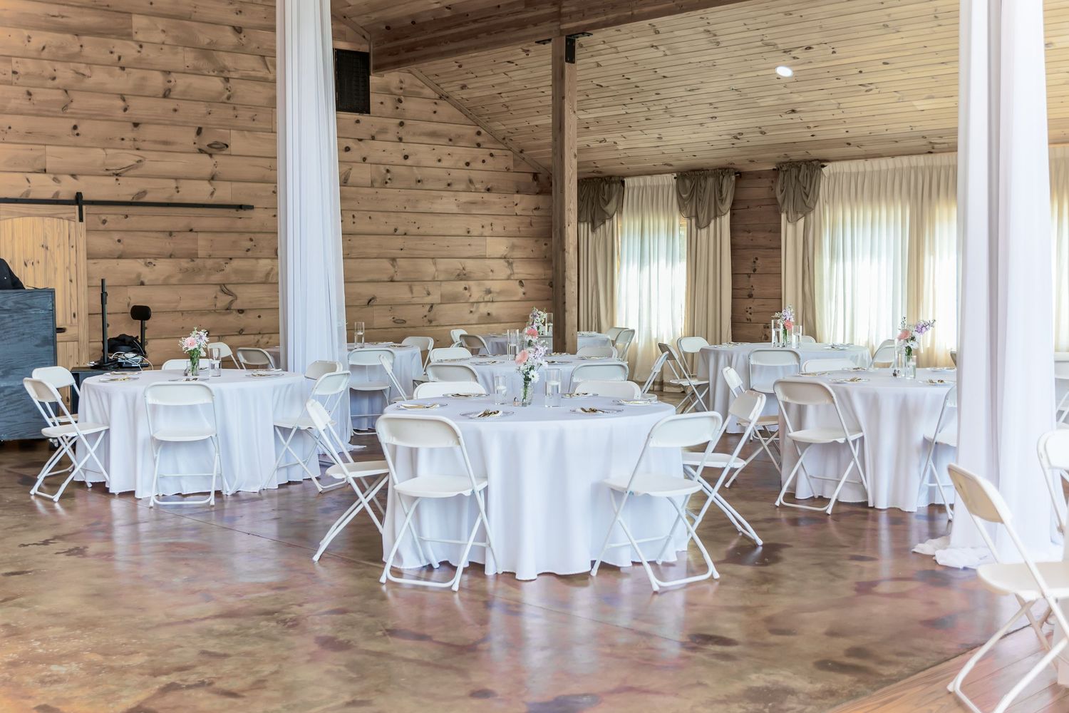 Rustic wedding reception venue with white round tables and folding chairs on concrete floor.