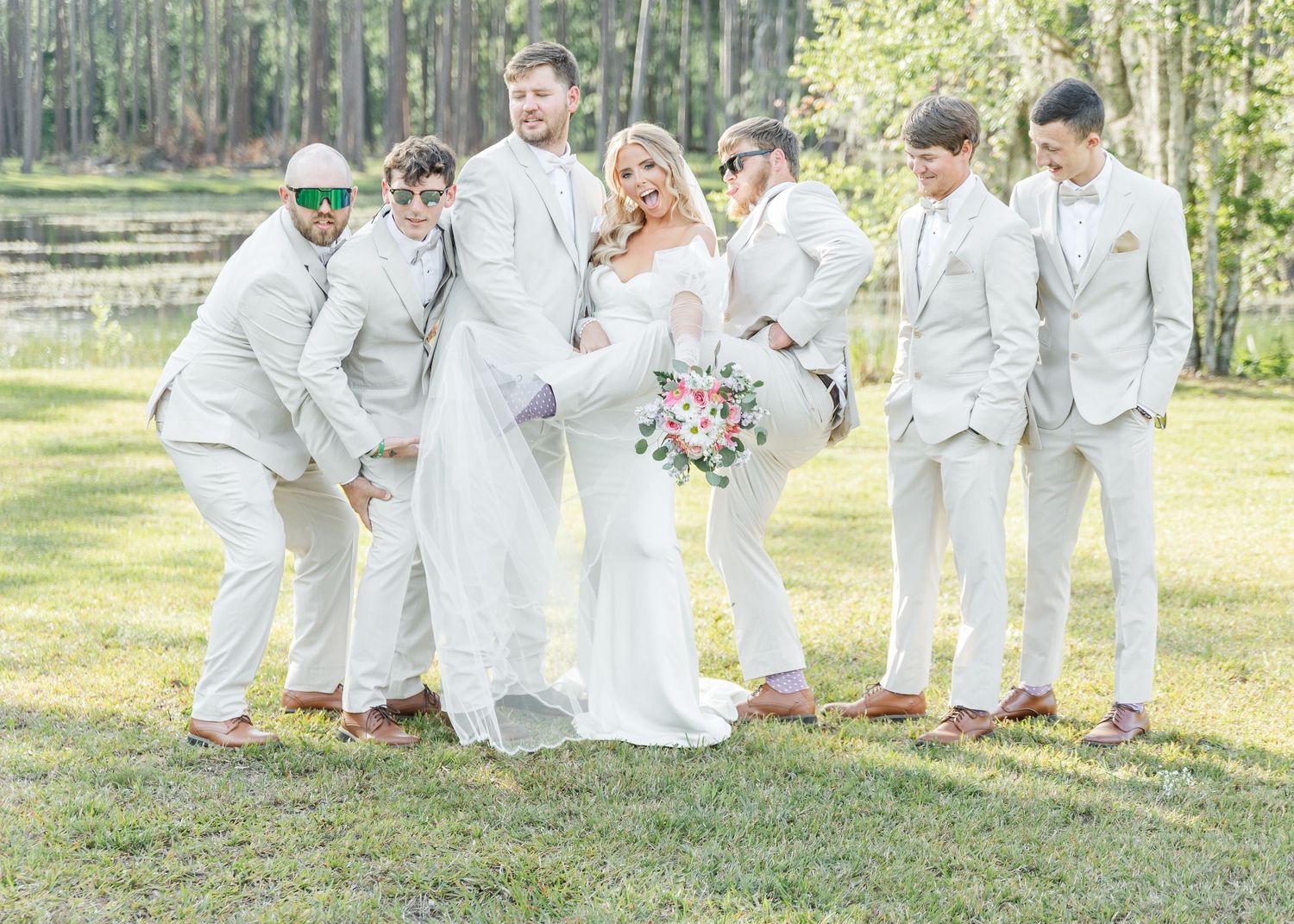 Groomsmen in white suits strike playful poses with couple outdoors.