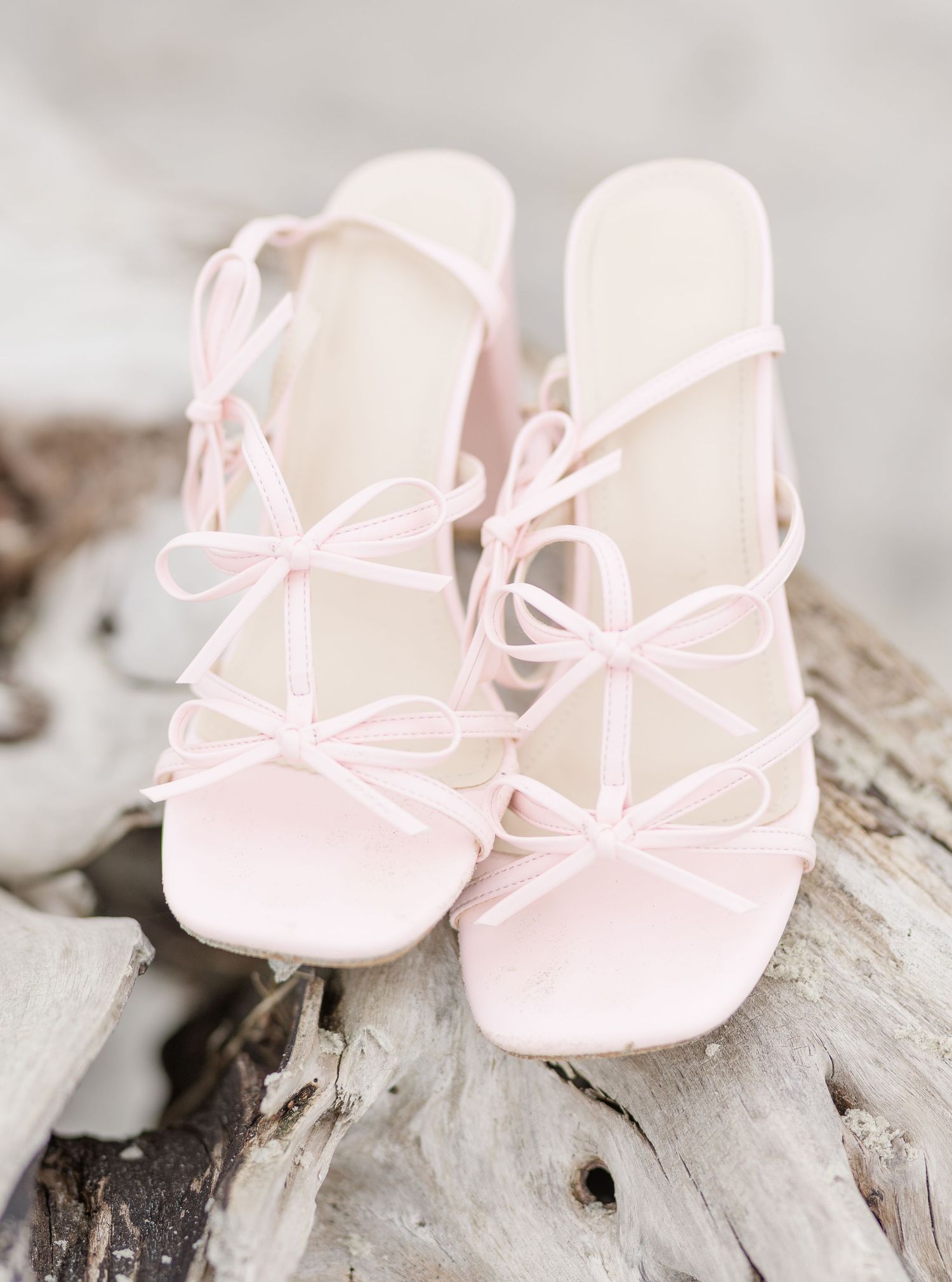 Delicate pink strappy sandals with bow details rest on weathered driftwood.