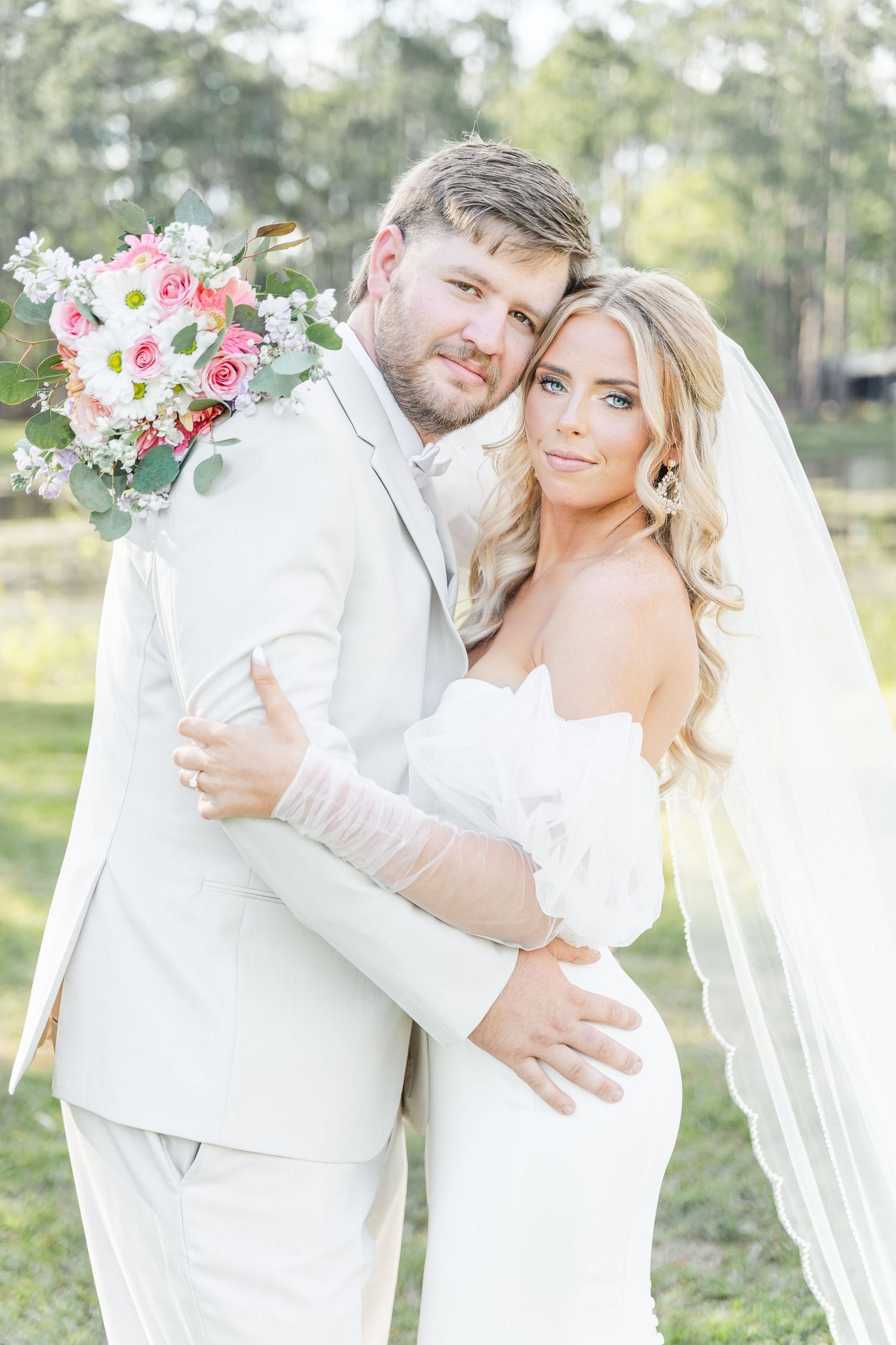 Wedding couple poses together in white attire holding a pink and white floral bouquet.