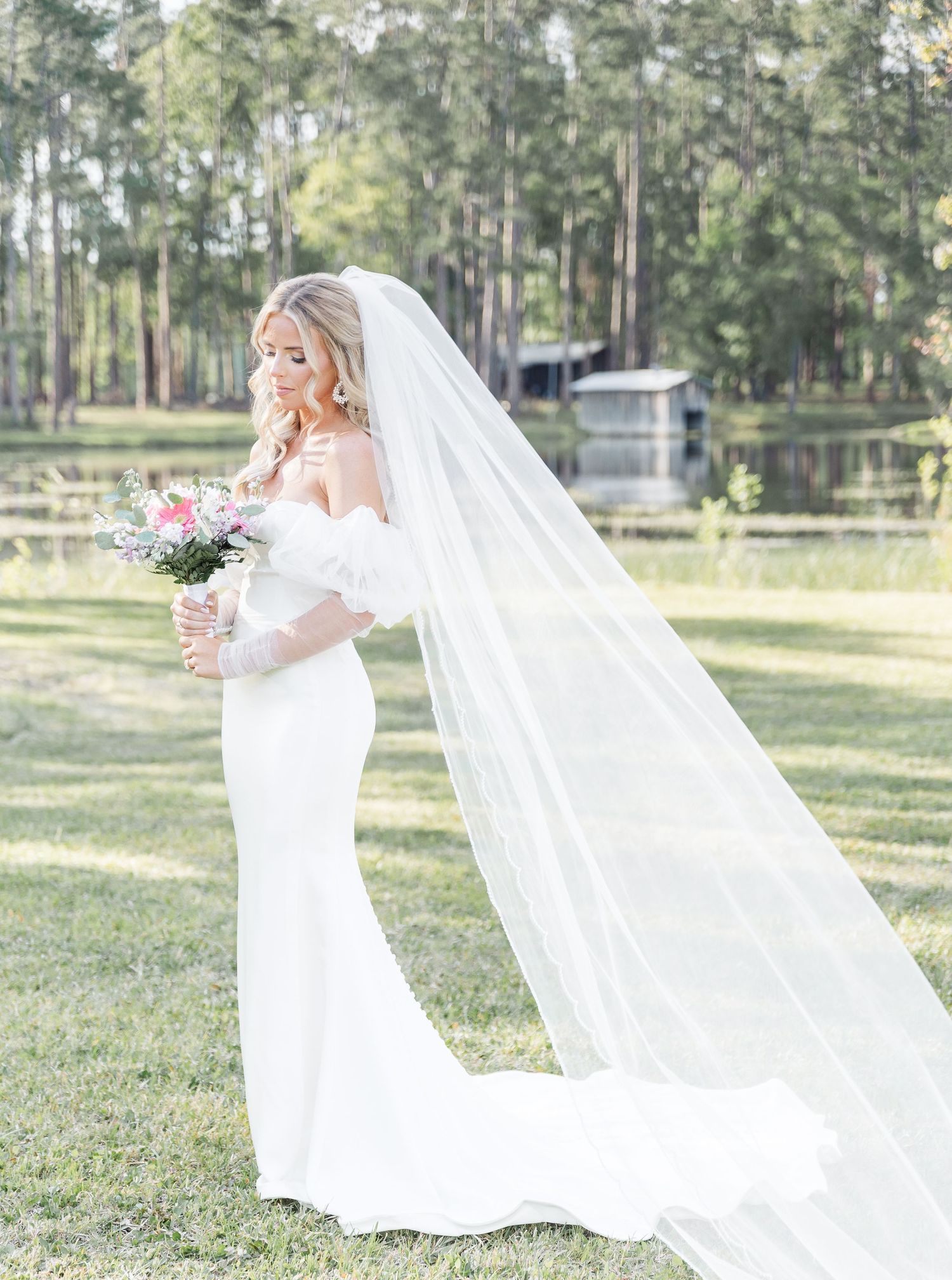 Bride in fitted white gown with cathedral veil poses by lake with flowing train.