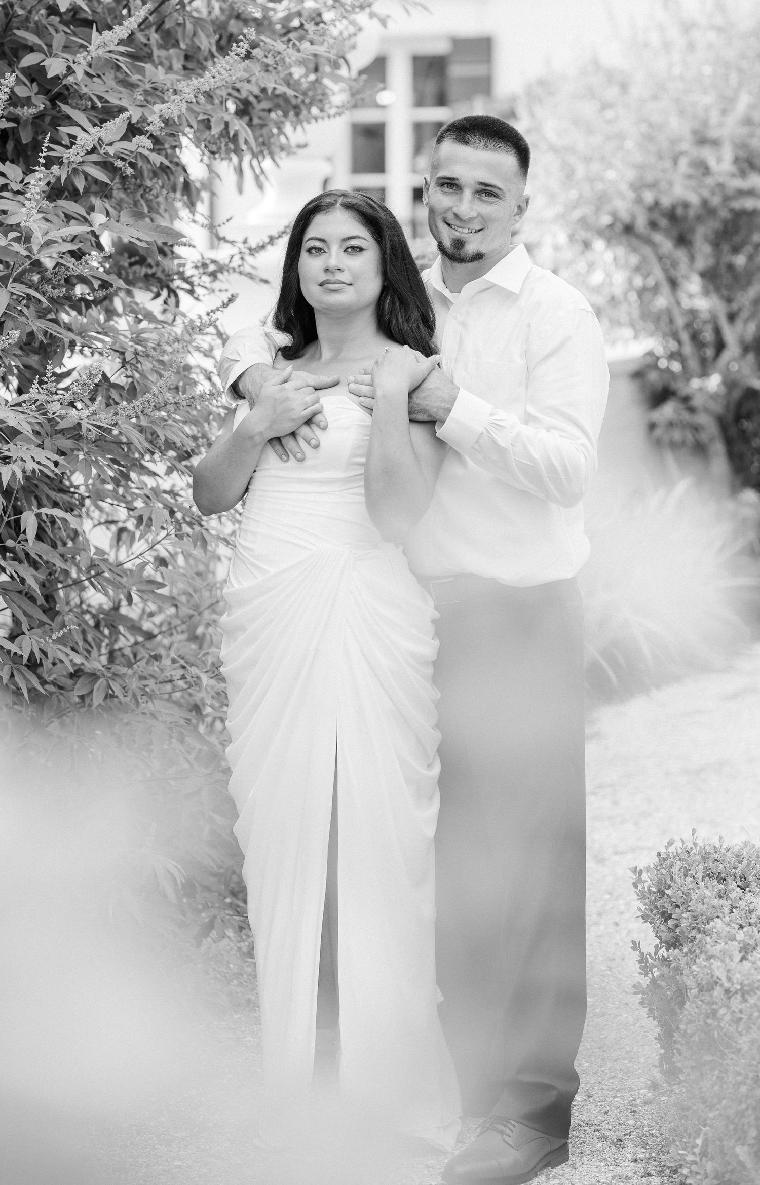 Black and white photo of a couple posing in a garden, bride in white gown with groom in white shirt behind her.