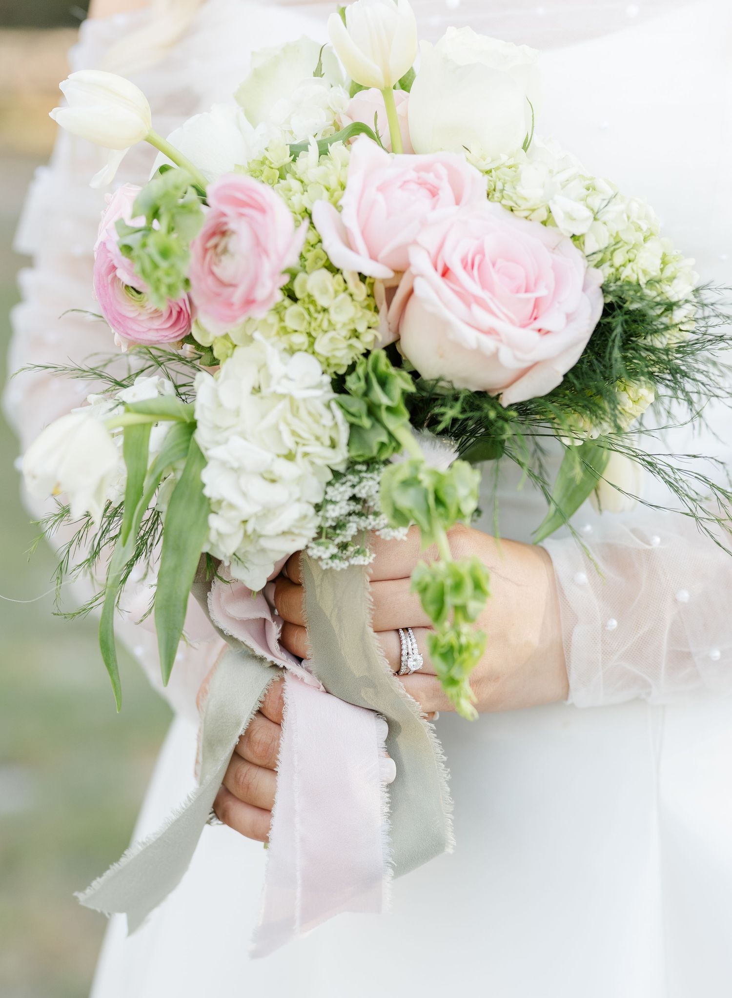 Bride holding a romantic wedding bouquet with pink roses, ranunculus, white hydrangeas, and greenery tied with a blush ribbon.