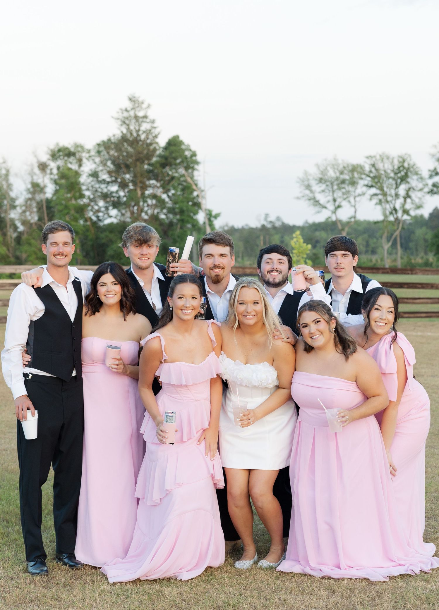 Wedding party posing outdoors in pink bridesmaid gowns and formal attire, smiling together on a green lawn.
