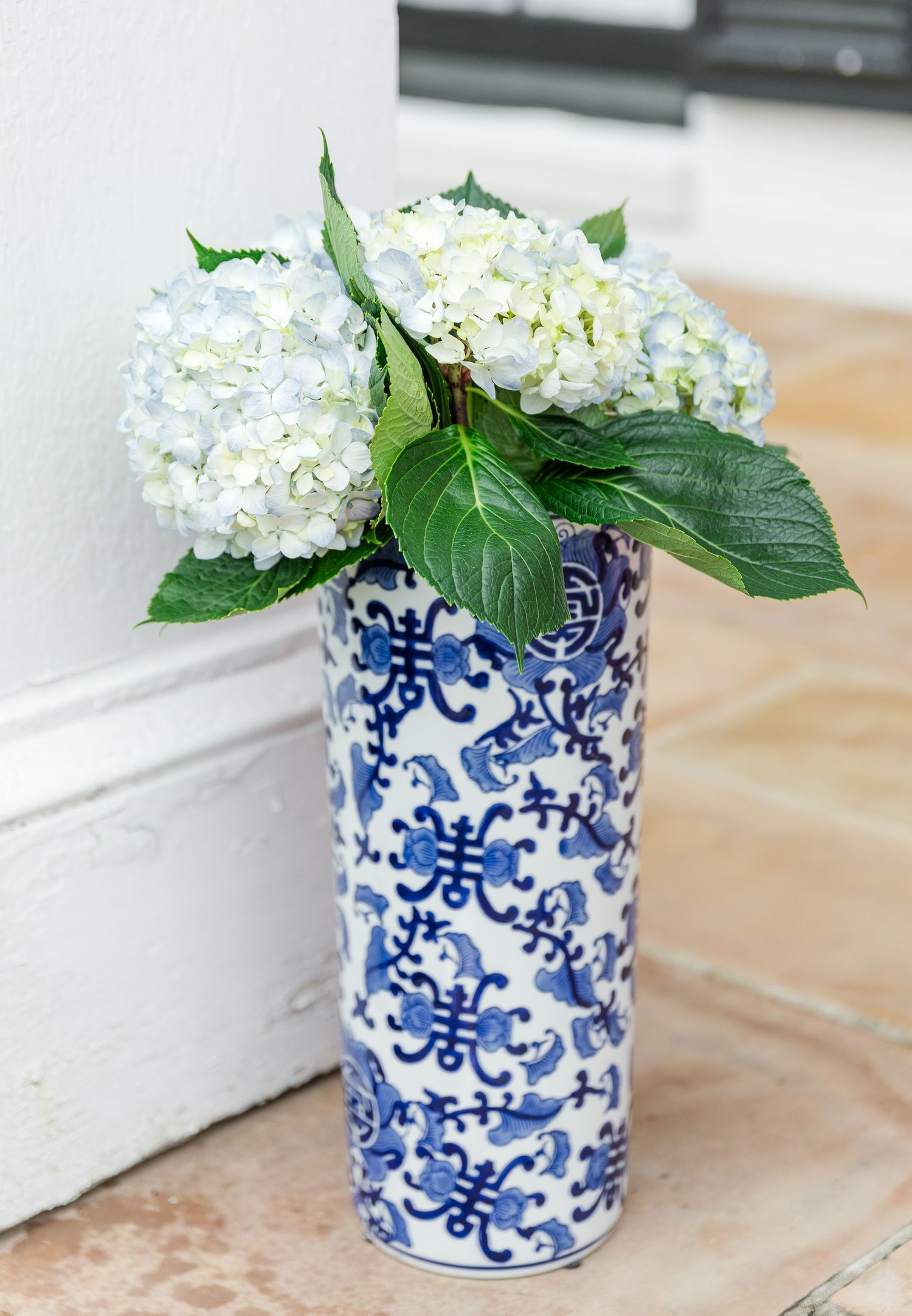 Blue and white chinoiserie vase with white and blue hydrangeas and green leaves on a tiled surface.