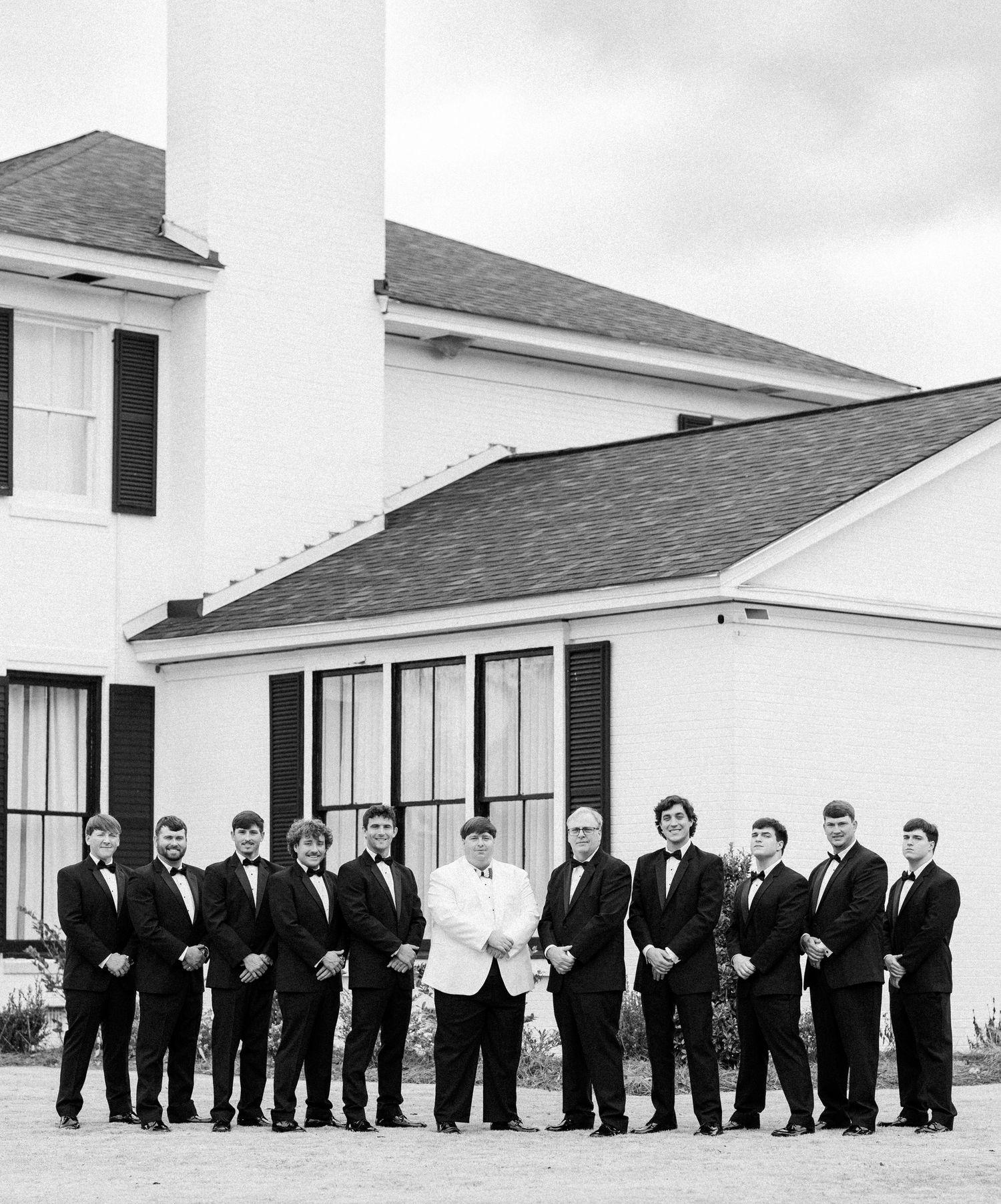 Black and white photo of groom in white jacket with groomsmen in black tuxedos outside white building.