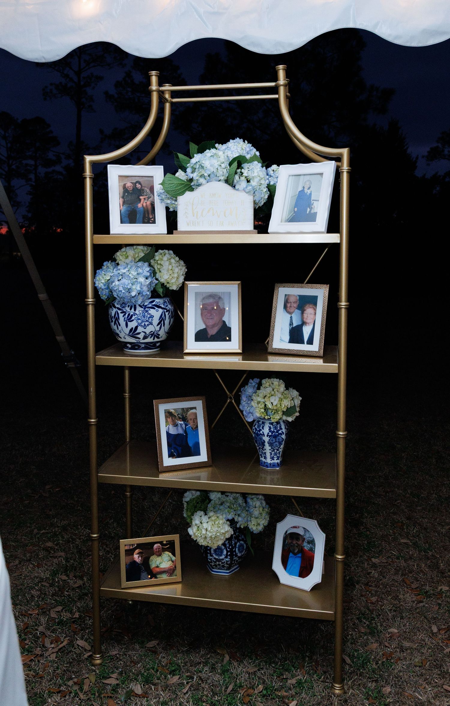 Gold bookshelf displaying framed family photos and blue-and-white chinoiserie vases with white hydrangeas at an outdoor wedding reception.
