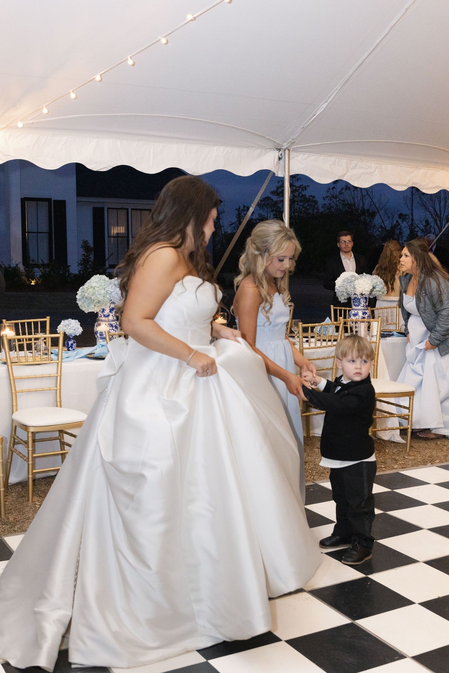 Bride in white ball gown dances with young boy in tuxedo on checkered floor at outdoor wedding reception.