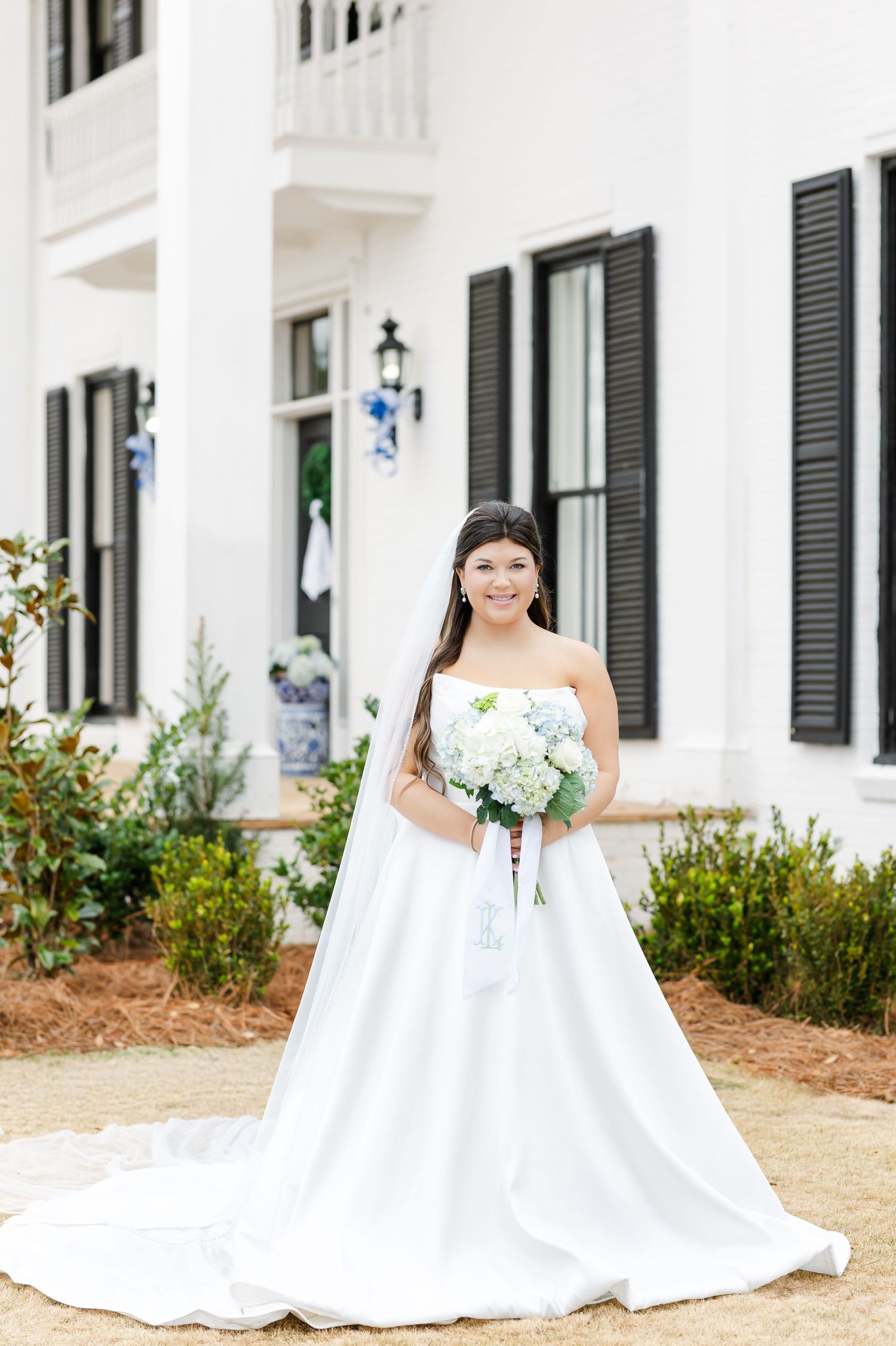 Bride in strapless white ball gown holds white hydrangea bouquet while posing in front of a white estate.