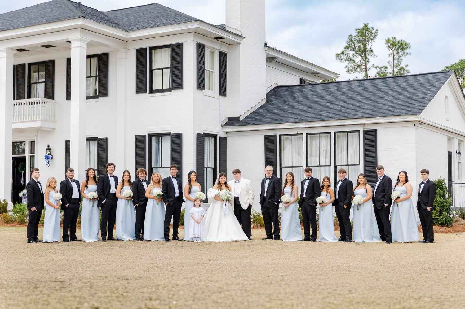 Large wedding party posing in front of a white Southern mansion with bridesmaids in light blue gowns and groomsmen in black tuxedos.