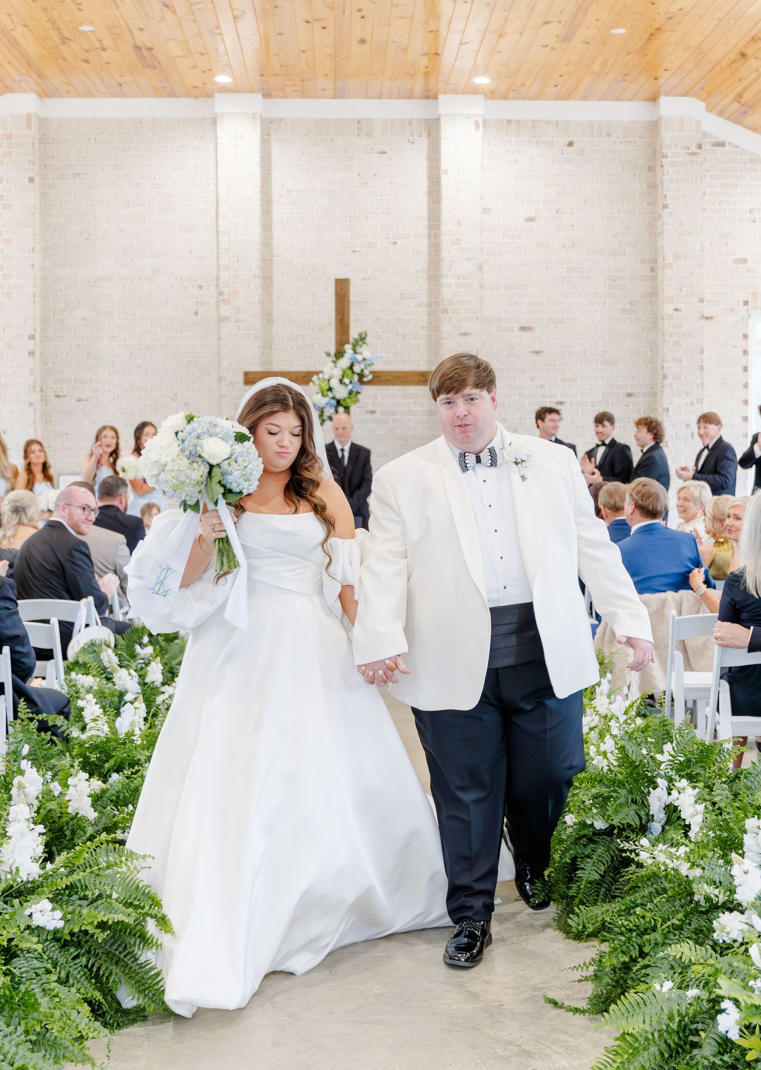 Newlyweds walk down the aisle lined with lush greenery after their ceremony in a modern chapel.
