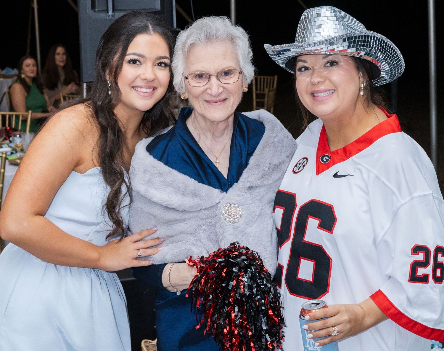 Bridesmaid in blue dress poses with elderly woman and guest wearing Georgia Bulldogs jersey at reception.