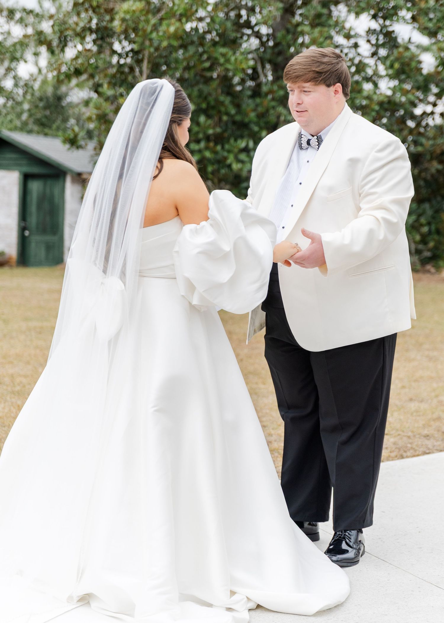 Groom in white tuxedo jacket holds bride's hands during outdoor first look wedding moment.
