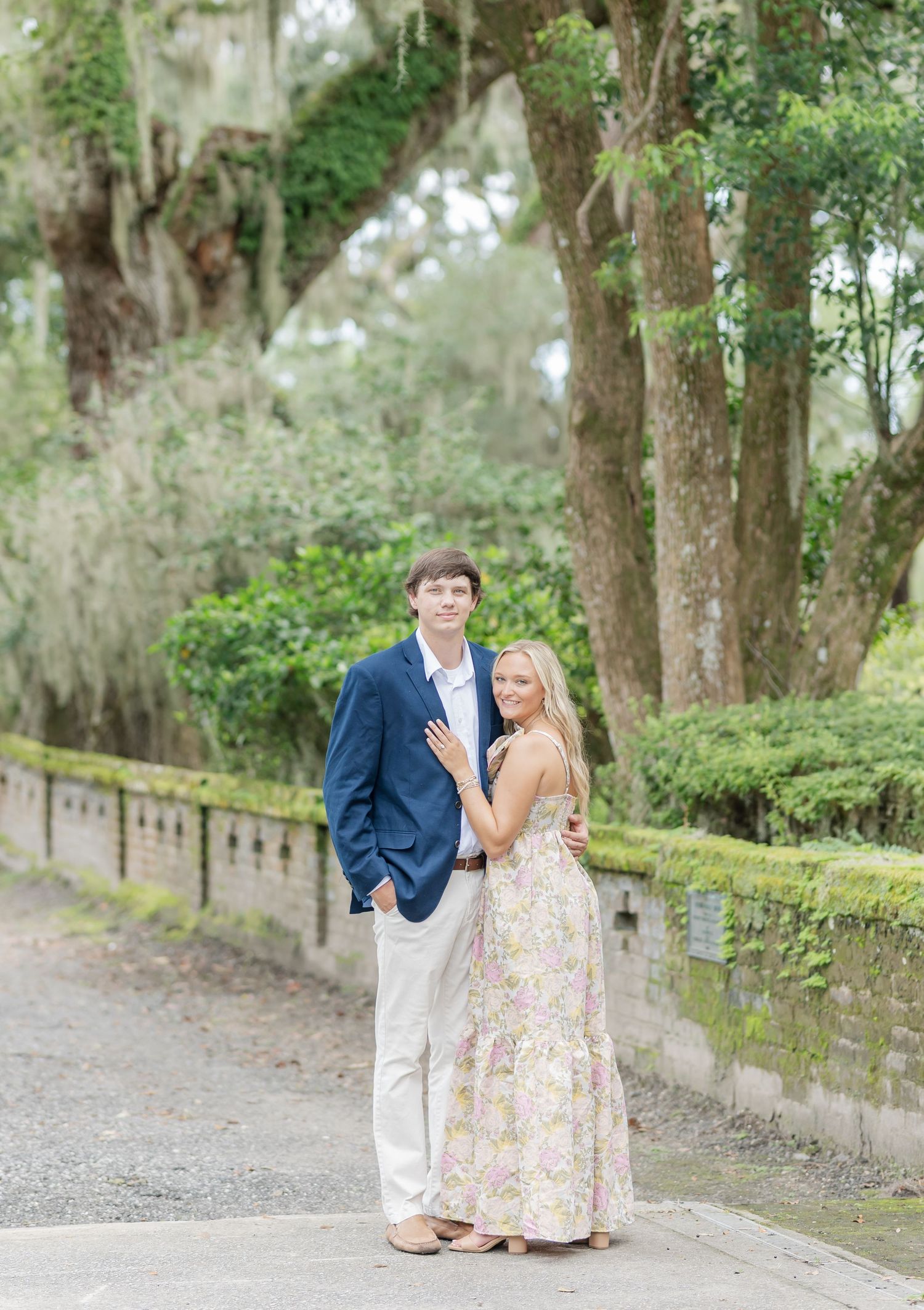 Couple poses romantically on garden path beneath moss-draped trees in springtime setting.