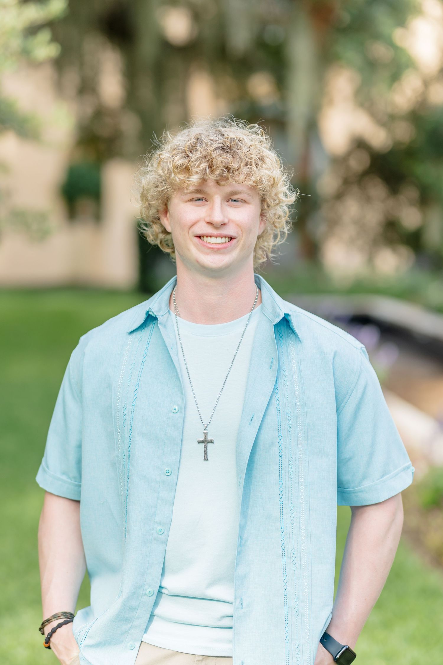 A casual outdoor portrait featuring someone in a blue button-down shirt over a white t-shirt with khaki pants.
