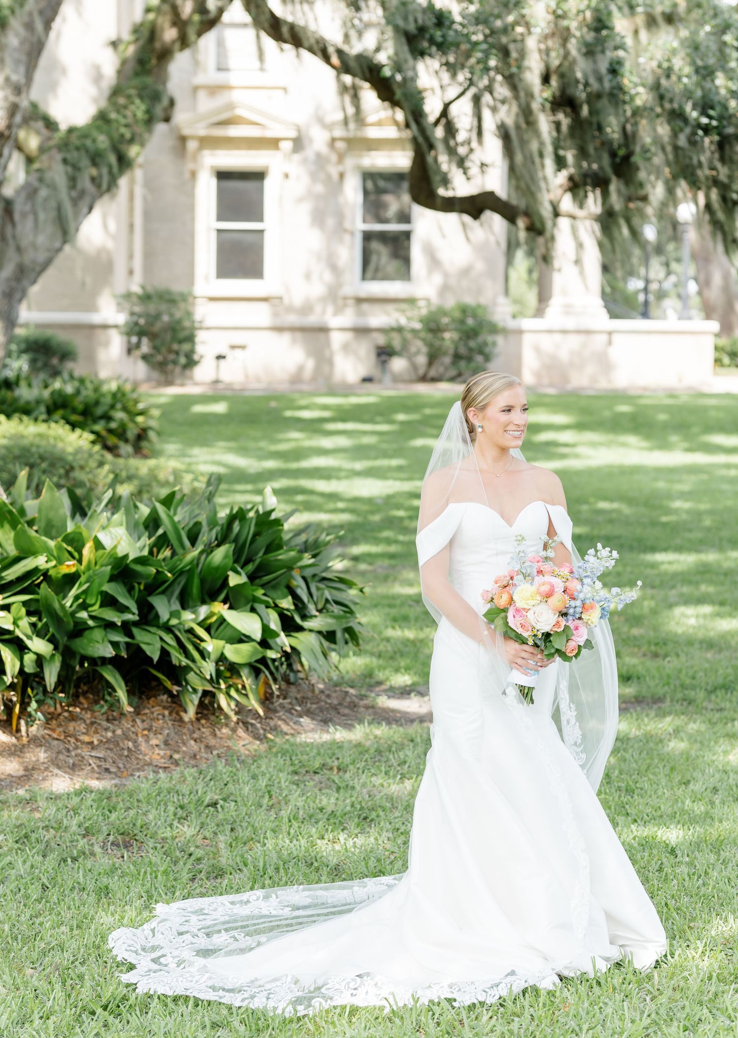 A bride in an off-shoulder wedding gown poses with bouquet on a manicured lawn near a historic building.