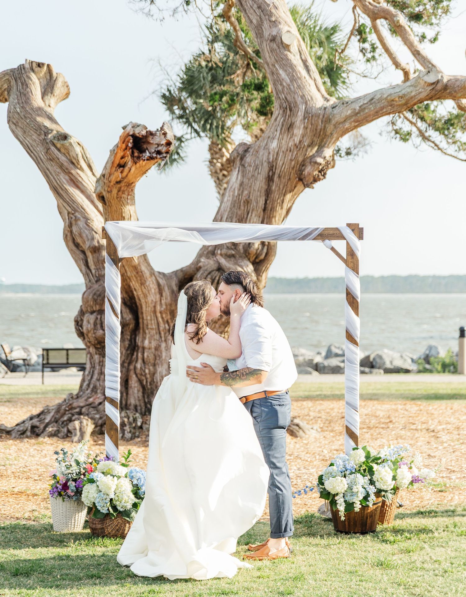 A romantic seaside wedding photoshoot featuring a couple posing under a gnarled tree on a sandy beach.