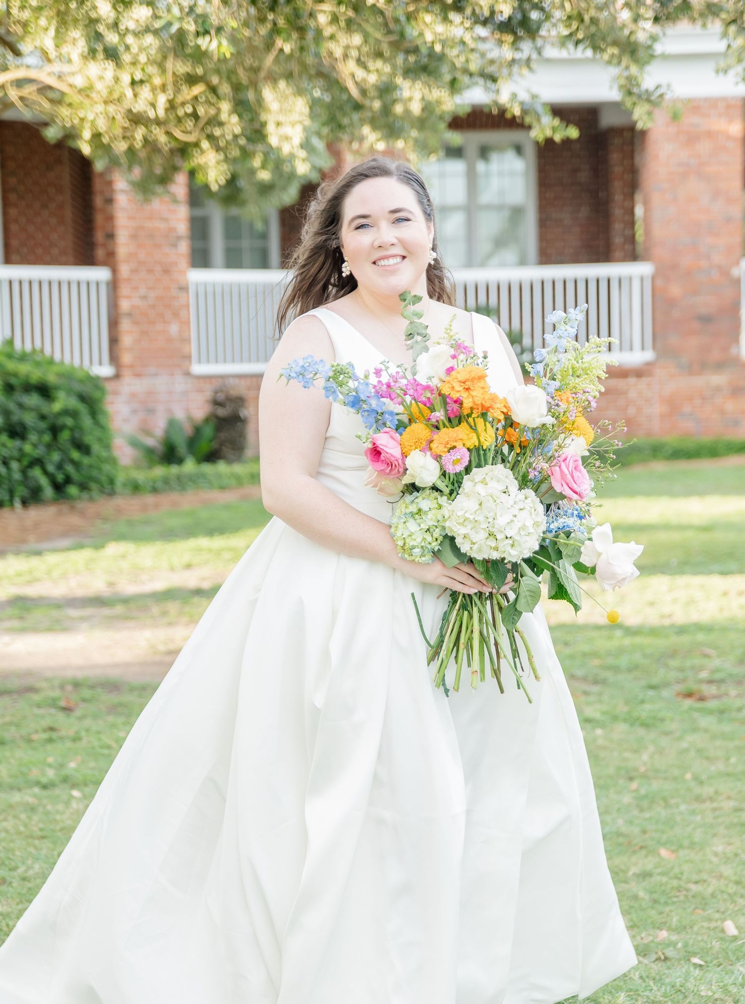 A bride in a flowing white gown holds a vibrant bouquet of colorful flowers while standing on a lawn near brick buildings.