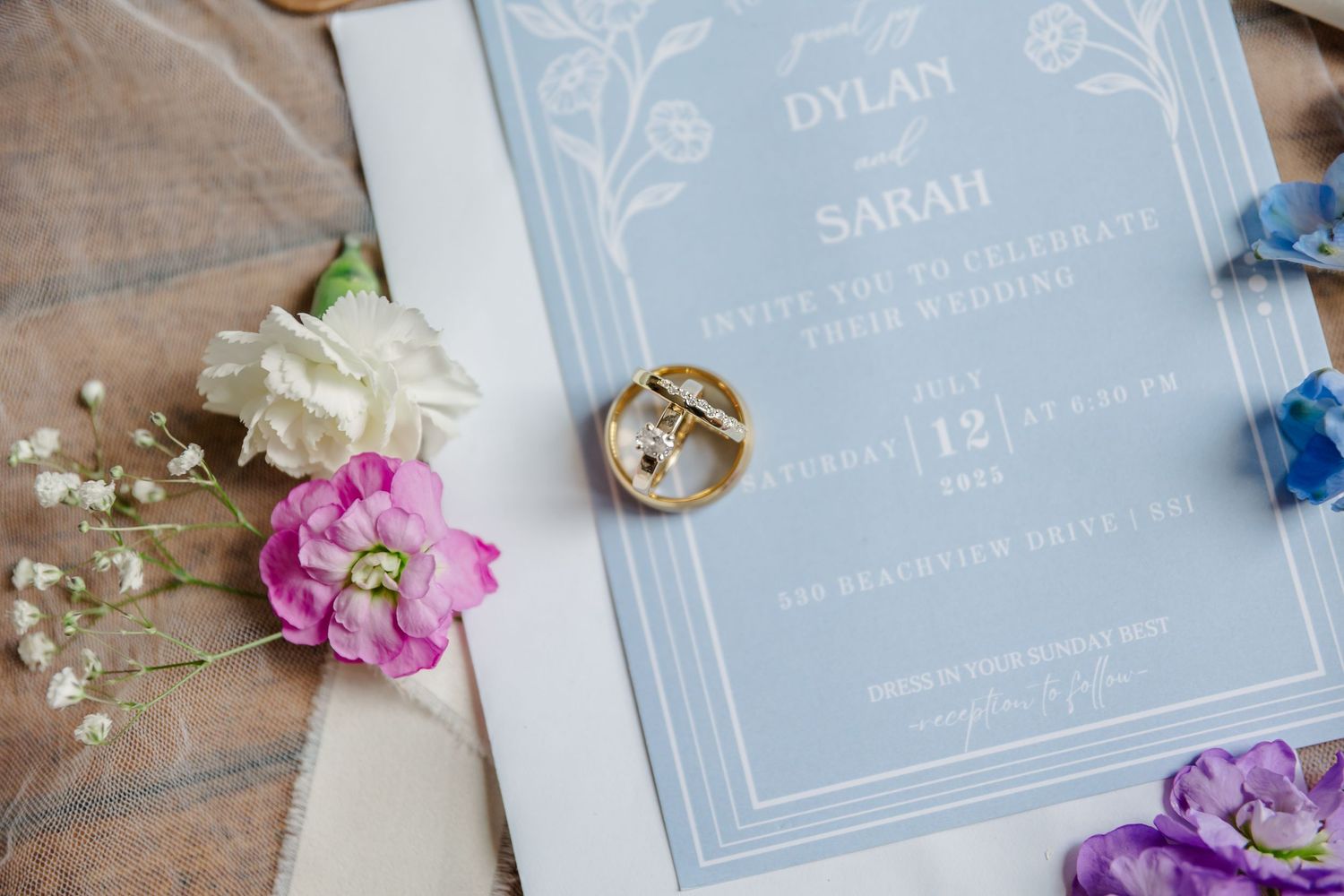 Wedding invitation suite in soft blue and white with floral accents, rings, and delicate baby's breath flowers on wooden surface.