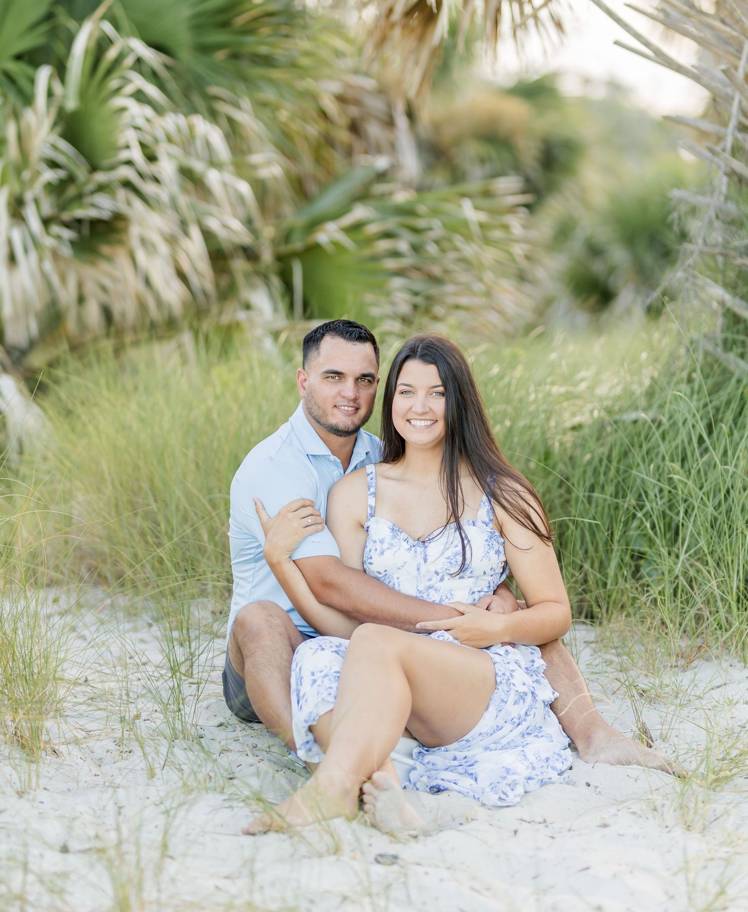 Couple sitting together on a sandy beach with palm trees in the background.