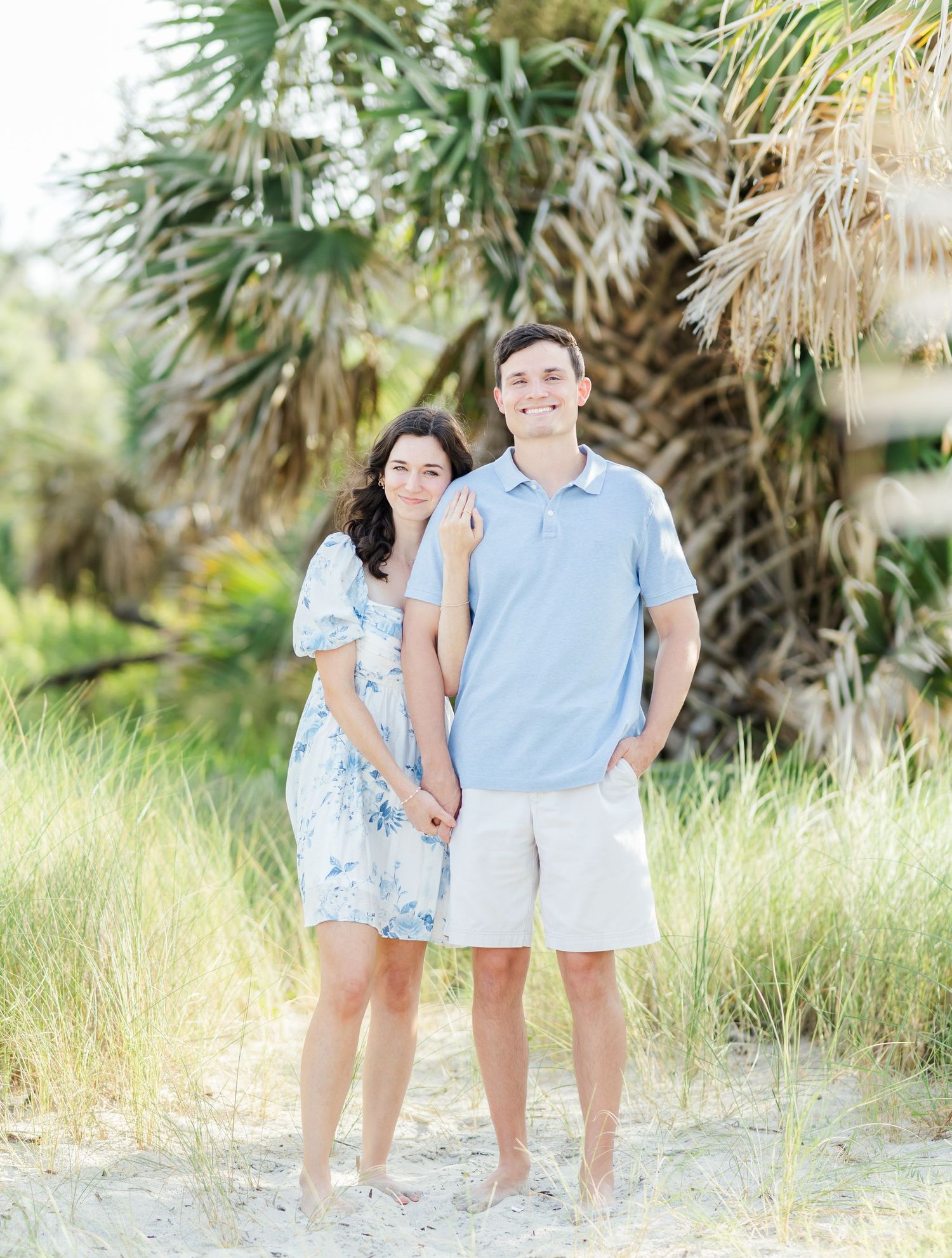 Two people in light blue and white outfits stroll together on sandy beach path near tropical palm trees.
