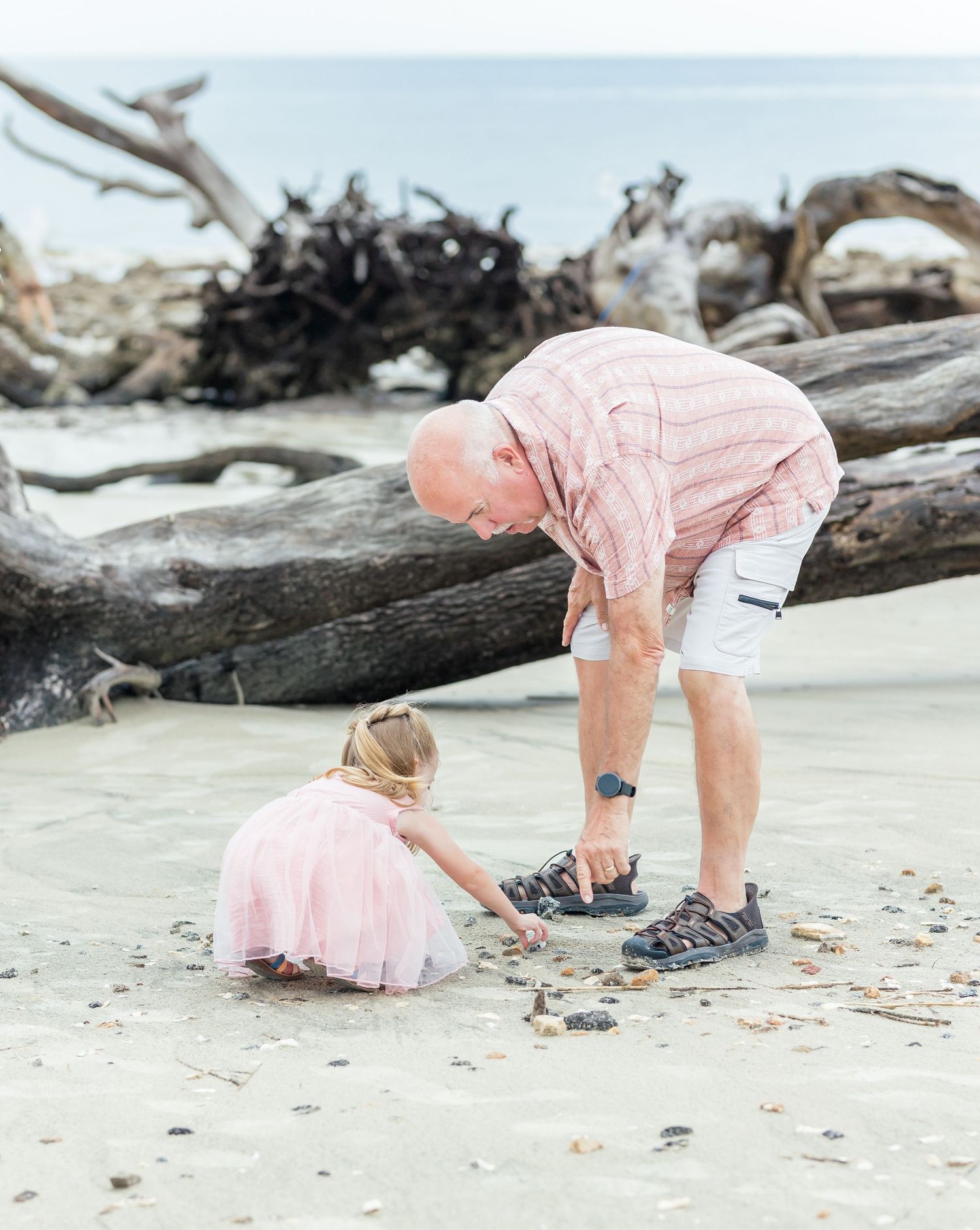 Two people explore and collect shells together on a beach with driftwood, both wearing pink outfits.