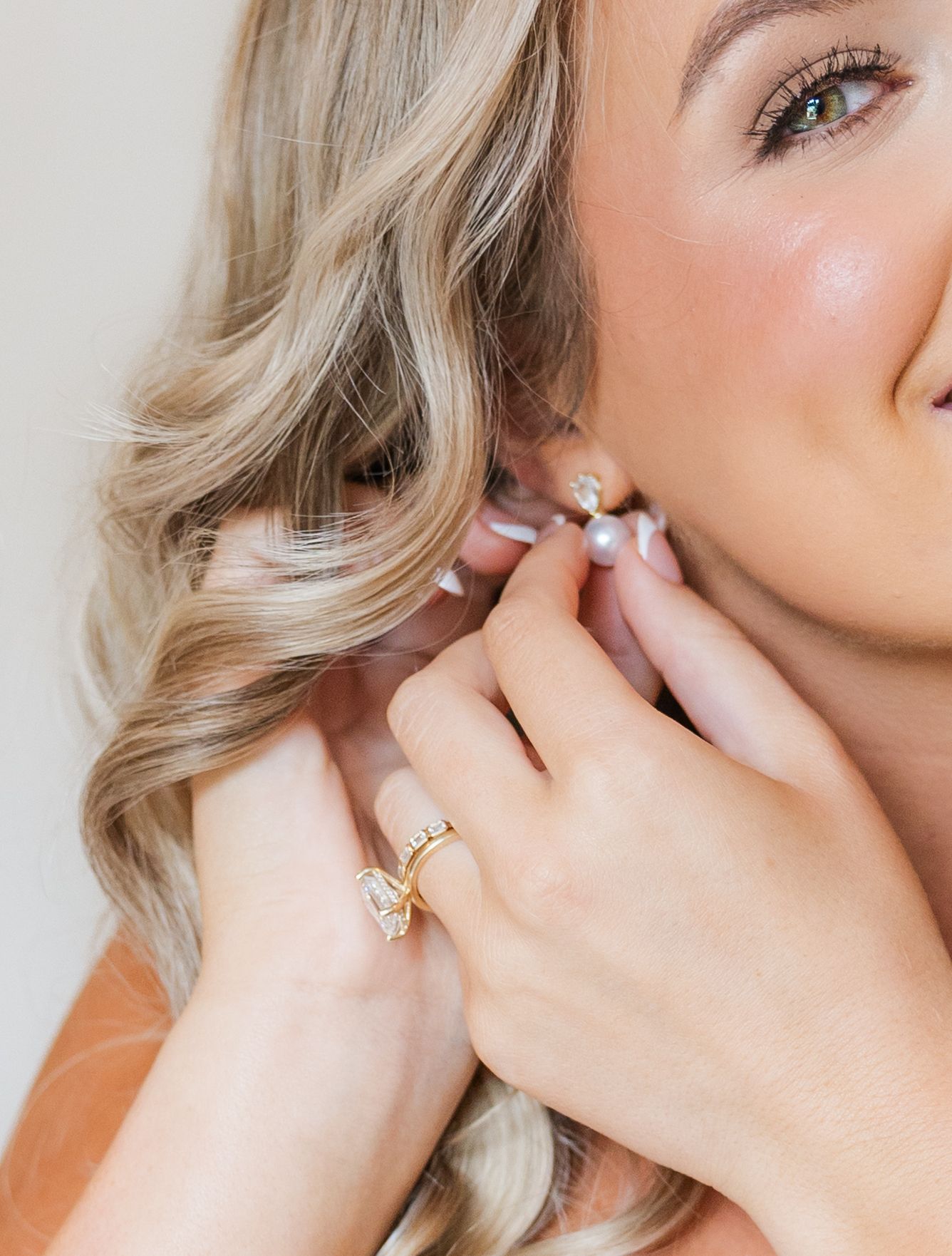 Blonde woman putting on a pearl earring, wearing a gold initial ring, with soft wavy hair and natural glam makeup.