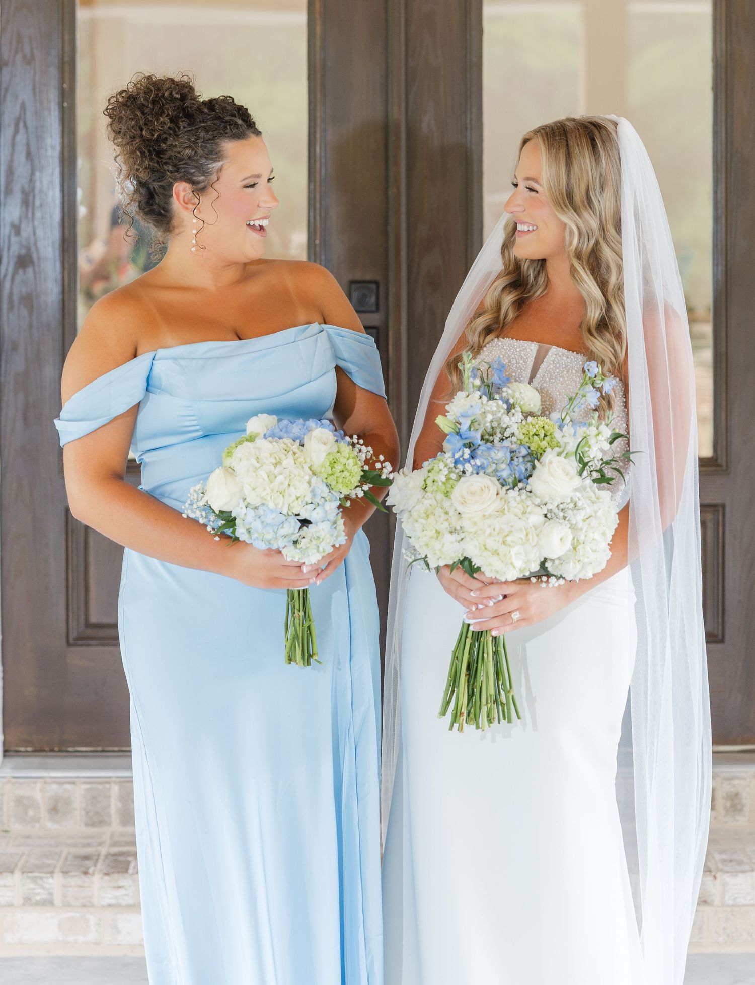 Bride in white gown and bridesmaid in light blue off-shoulder dress holding floral bouquets.