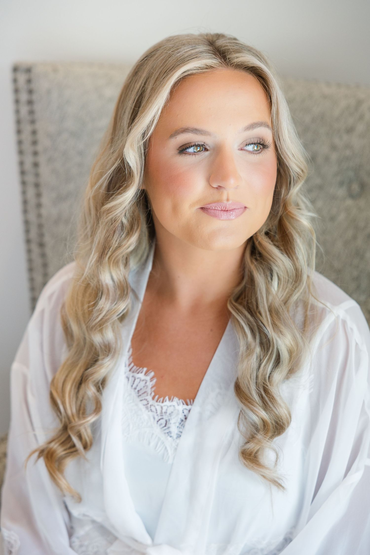Bride with long blonde wavy hair wearing a white lace robe, showcasing elegant bridal makeup.