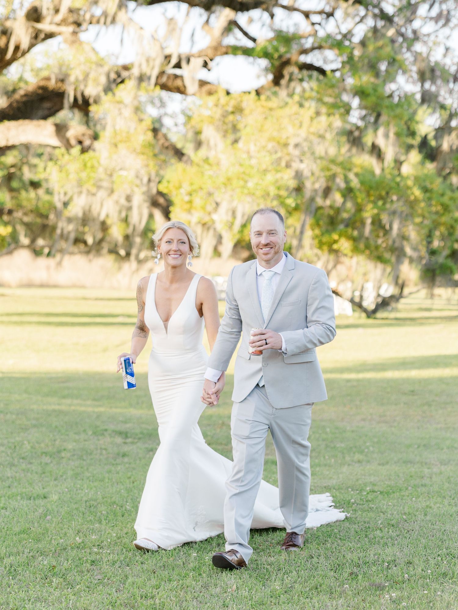 Bride in white gown and groom in gray suit walking hand in hand across a sunlit green lawn.