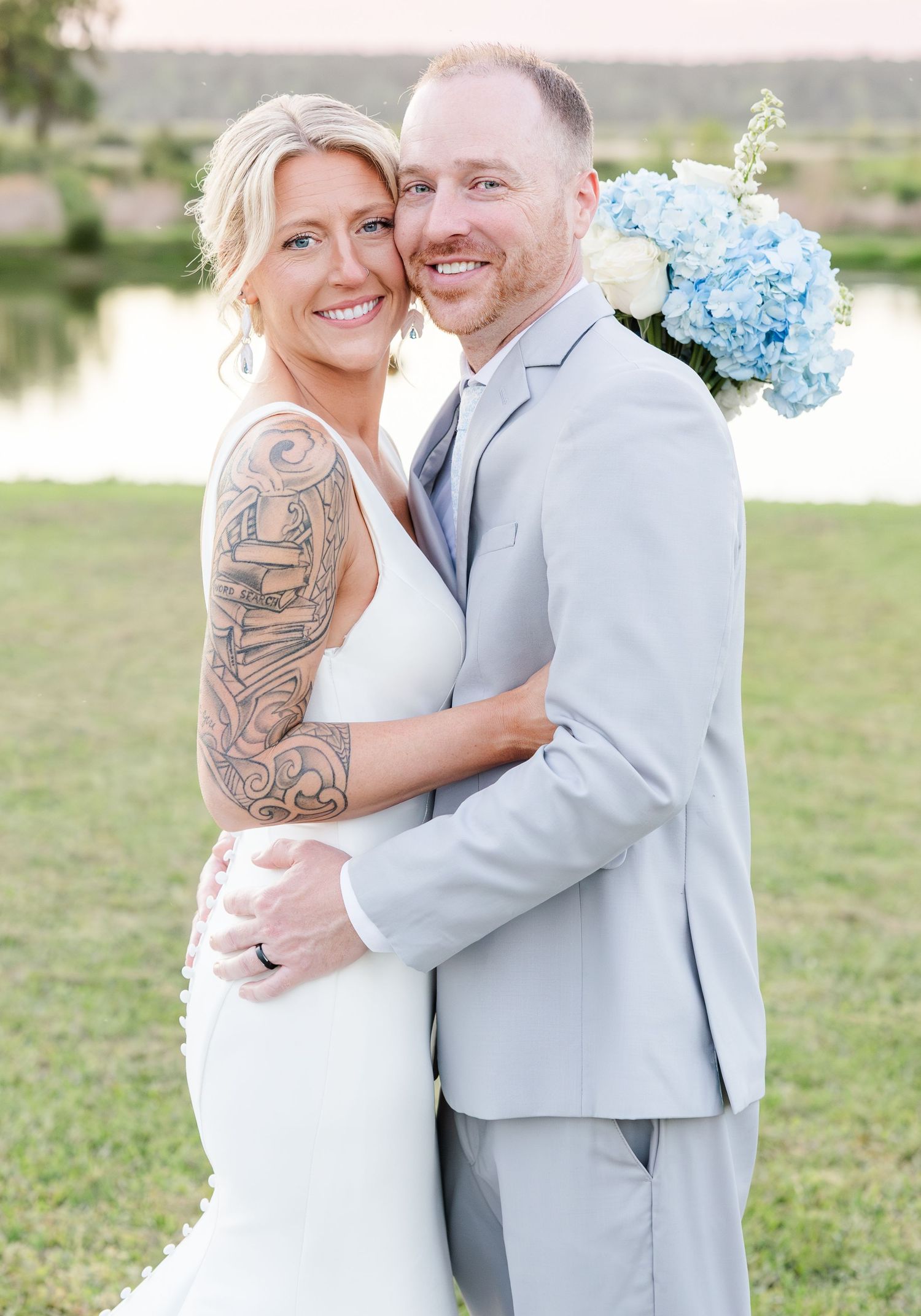 Smiling bride with sleeve tattoo and groom in gray suit embrace at outdoor wedding.